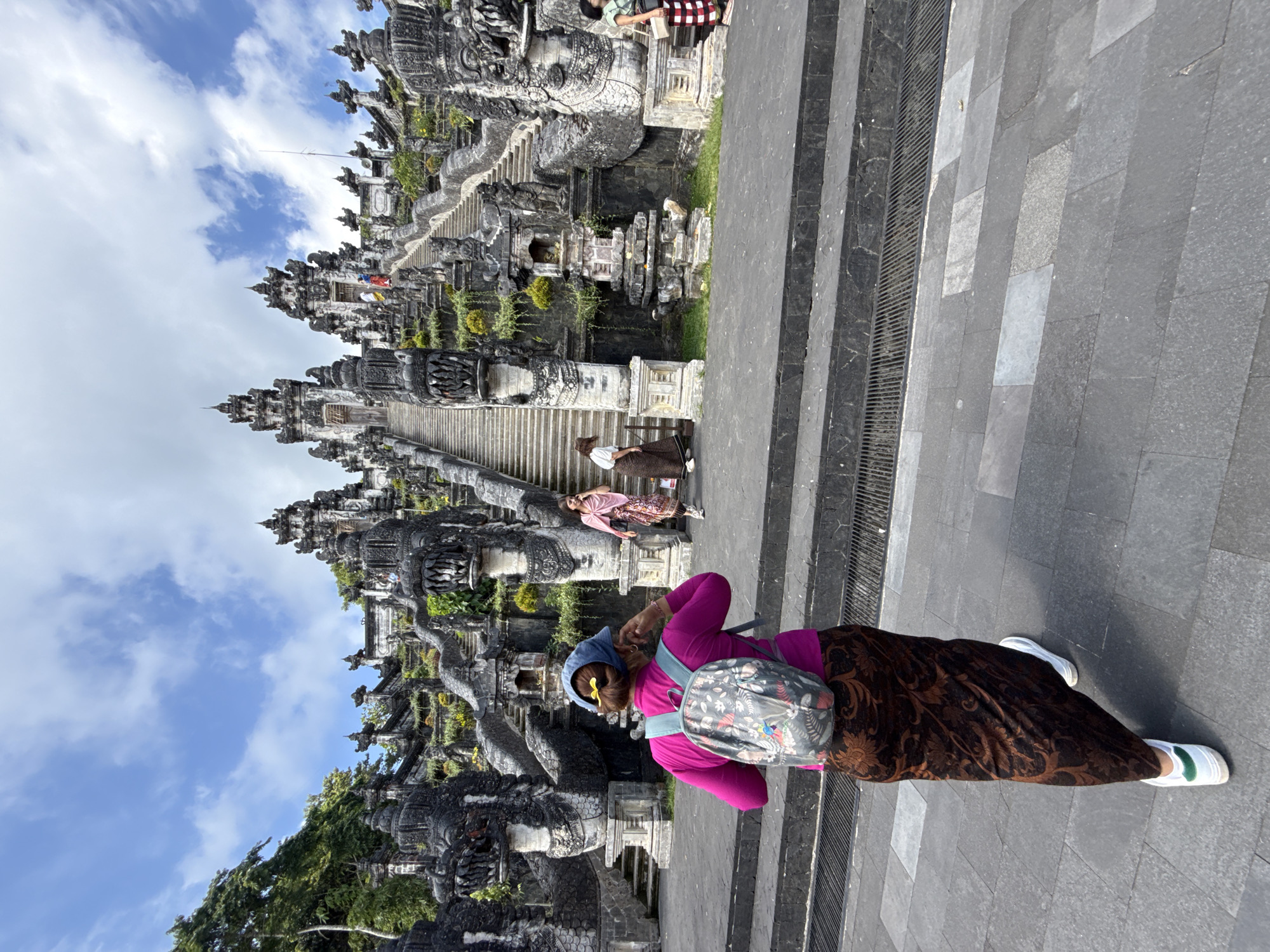 The image shows a wide shot of the Lempuyang Temple in Abang, Indonesia. The temple features a grand stone staircase with intricate carvings of dragon-like figures lining the sides. Several people are present, with one woman in the foreground wearing a magenta top and a patterned brown sarong and a backpack, facing away from the camera. Two other women are visible further up the stairs, one in a pink top and sarong, and another in a white top and brown sarong. The temple is adorned with lush greenery and elaborate architectural details under a bright blue sky with scattered clouds.