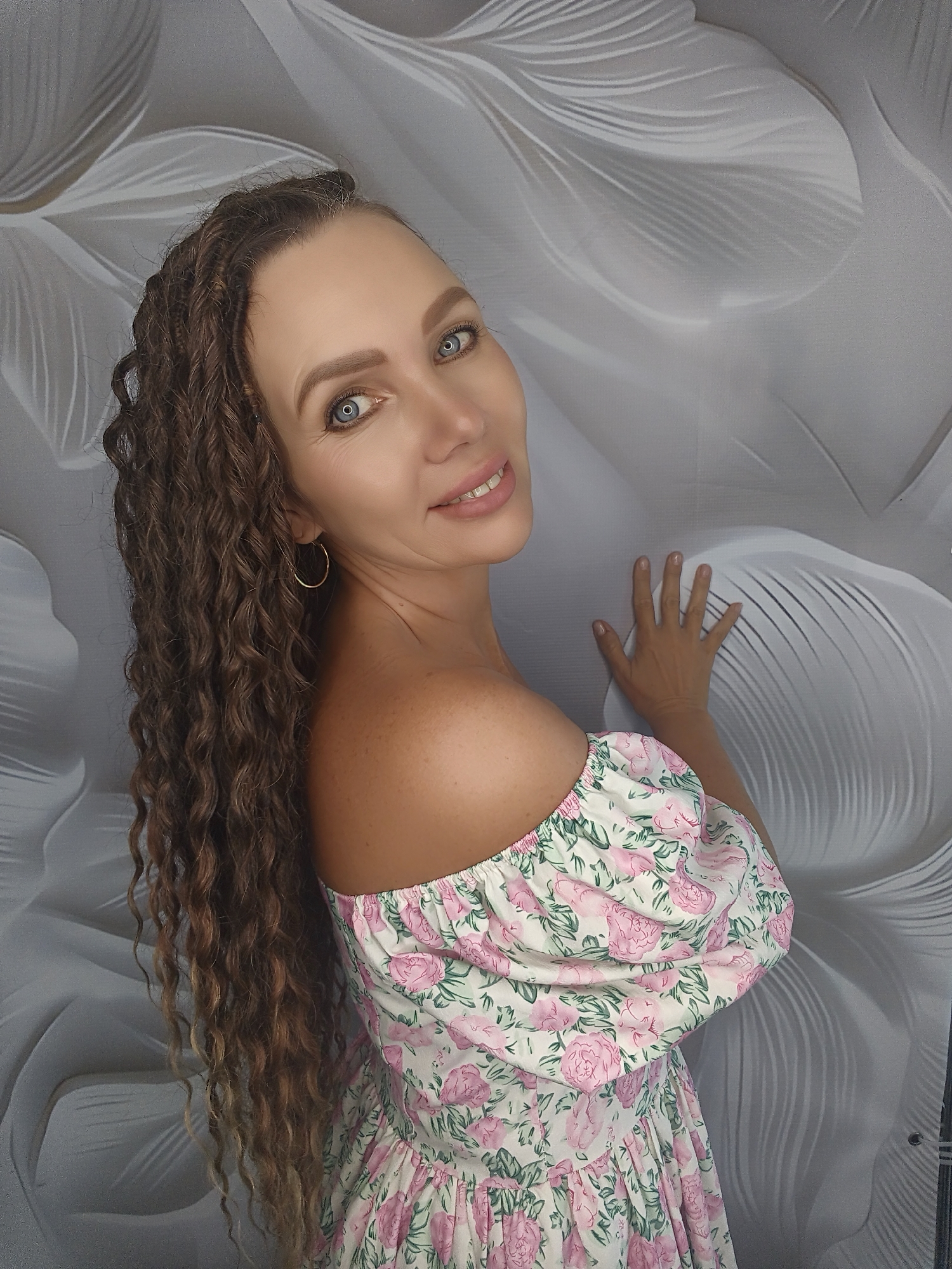 A woman with long, curly brown hair and blue eyes is shown from the chest up, angled to her left and looking over her left shoulder at the camera. She is wearing an off-the-shoulder dress with a floral print of pink roses and green leaves on a white background. Her left hand is resting on a grey, textured wall with a pattern of large, abstract floral shapes. She is smiling.