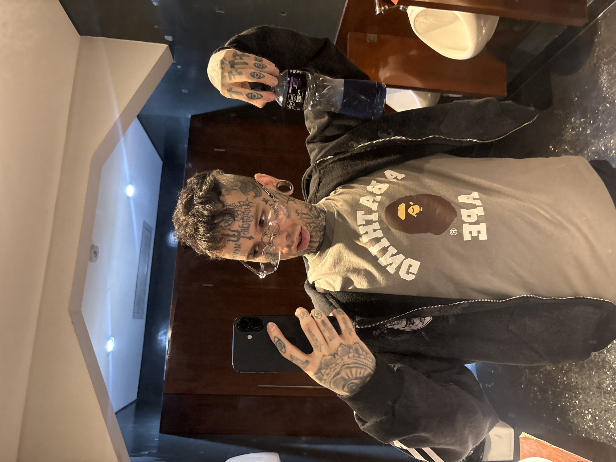 A male individual with extensive facial, neck, and hand tattoos is depicted taking a mirror selfie in what appears to be a men's restroom within a commercial or public establishment in Buenos Aires, Argentina. The exact location beyond a public restroom in Buenos Aires cannot be determined from the image.
The subject has short, curly dark hair and prominent black ear gauges. He wears clear, rimless glasses with intricate metallic detailing on the bridge and temples. His attire consists of a light grey t-shirt featuring a brown "Bathing Ape" (BAPE) logo and partially visible text, layered under an open dark grey or black zip-up hoodie with a white stripe visible on the left sleeve.
In his left hand, the individual holds a black smartphone, likely an iPhone model with dual rear cameras, pointed towards the mirror to capture his reflection. His right hand holds a partially full, clear plastic Gatorade bottle containing a purple liquid. The label is visible, confirming the brand.
The environment includes two white porcelain urinals on the right, separated by dark brown wooden partitions. The walls are dark, possibly black or dark grey, and appear somewhat textured or reflective. Dark brown wooden panels or doors are present in the background. The floor is dark and reflective, showing speckles of light. The ceiling is white with recessed circular lighting fixtures, one of which emits a bright white light.