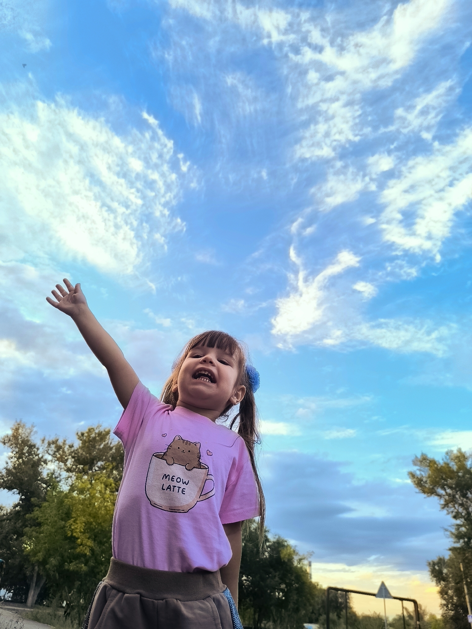 In Qarağandı city administration, Kazakhstan, a young girl with brown hair, styled in pigtails and adorned with a blue bow, stands outdoors against a bright blue sky filled with wispy white clouds. She is wearing a pink t-shirt featuring a graphic of a cat in a mug with the text "MEOW LATTE" and a brown skirt. Her right arm is raised, palm open, as she looks upwards with her mouth open, seemingly in mid-sentence or mid-scream. The background consists of green trees and what appears to be a fence or railing, suggesting a park or outdoor recreational area. The perspective is from a low angle, emphasizing the sky and the girl's joyful or energetic pose.