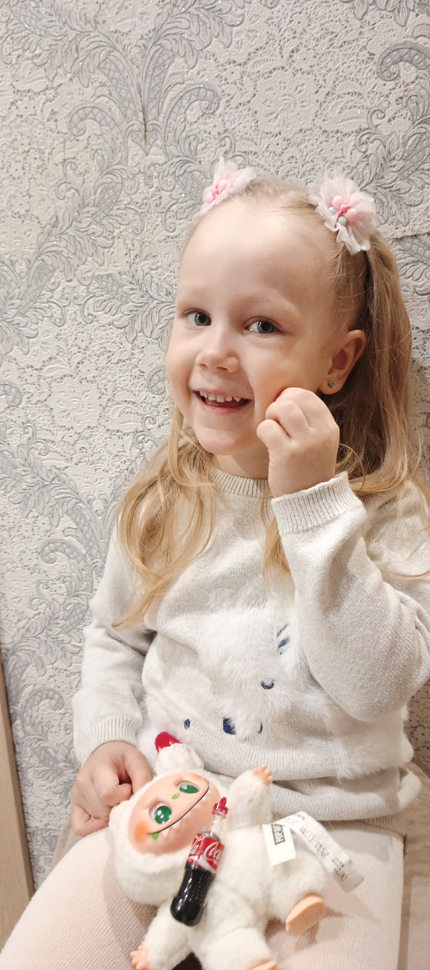 A young, fair-skinned child with blonde hair tied in pigtails adorned with pink floral hair accessories is smiling at the camera. She is wearing a light-colored sweater with a fuzzy pattern. Her right hand is touching her right cheek. A plush toy, possibly a Yeti, is nestled in her lap holding a miniature Coca-Cola bottle. The background is a patterned, possibly wallpapered, wall. The image was taken in the city of Chaikovsky, Russia.