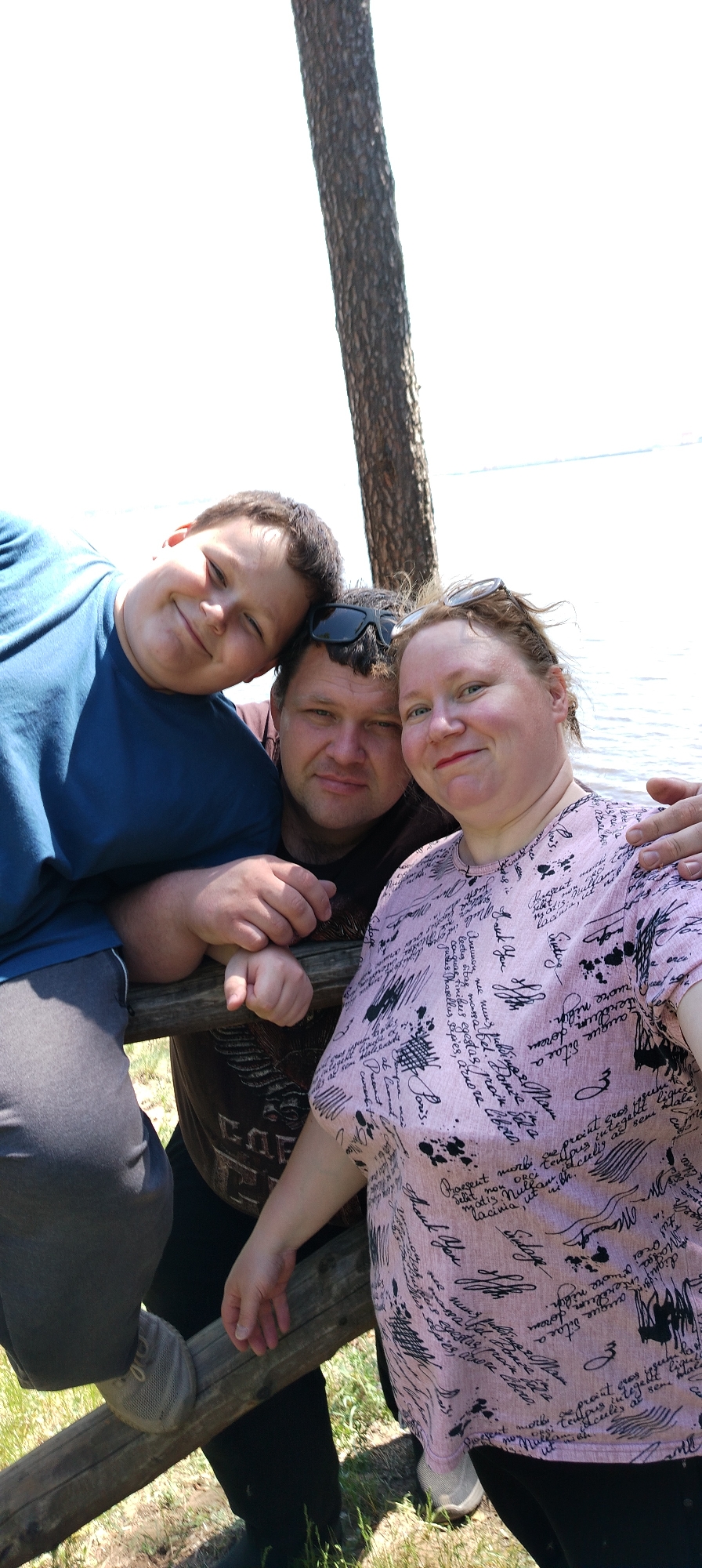 Here's a concise description of the image:
The photograph shows three people - a woman, a man, and a younger person - posing for a selfie outdoors. The man is in the center, with the woman to his right and the younger person to his left. They are leaning against a wooden fence or railing. Behind them, there is a tree trunk and a body of water in the background, suggesting a lakeside location. Based on the information provided the city where the photograph was taken is Noviy, Russia.