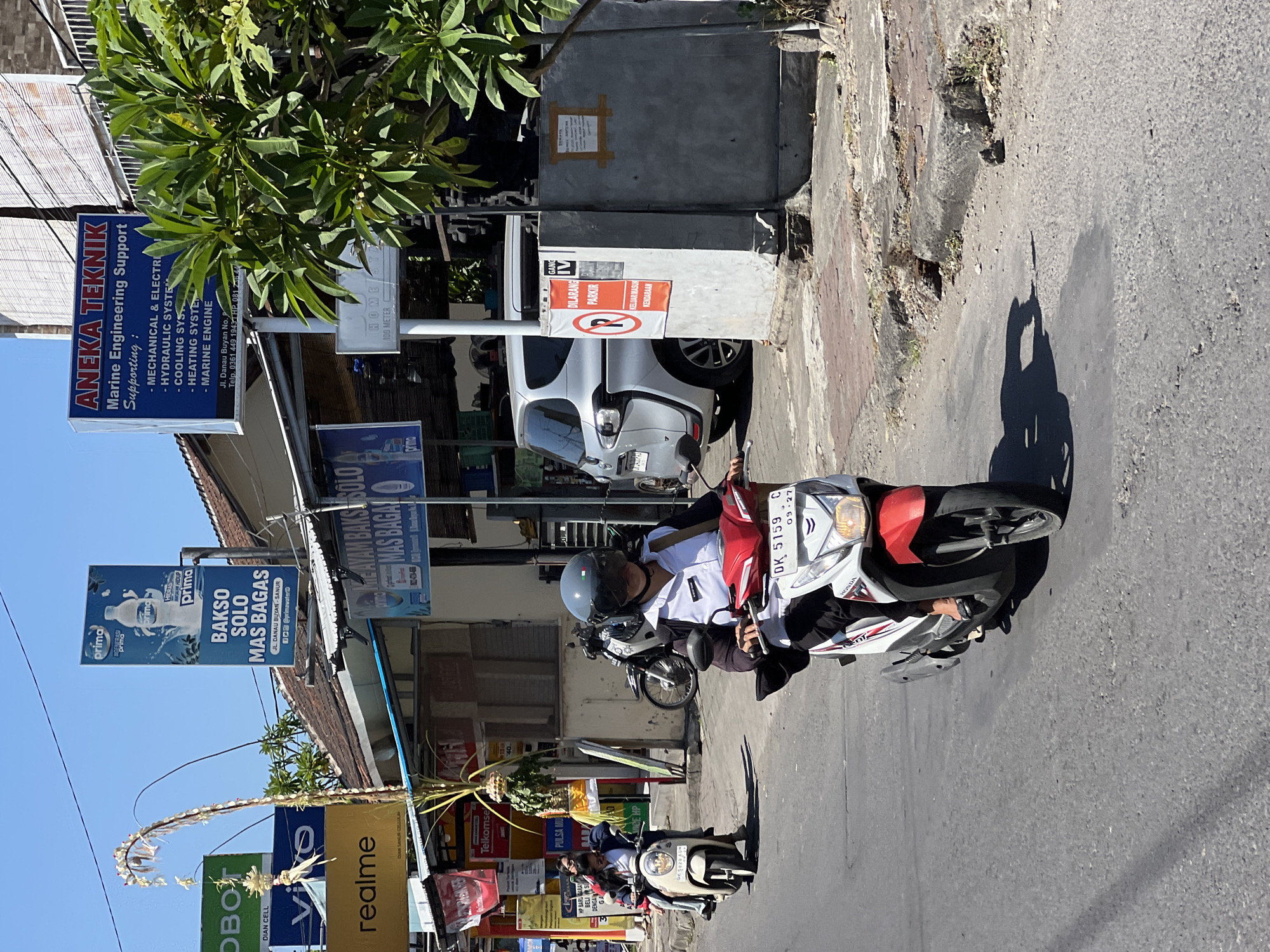 This is a street-level, daytime, medium shot photograph taken in Denpasar, Indonesia, showing a street scene with several motorcycles and commercial signage. In the foreground, a person wearing a helmet and a white collared shirt with dark sleeves is riding a red and white Honda scooter, license plate DK 5159 C. The scooter has a white front fender and red accents on the sides. Behind this scooter, another scooter with two passengers, both wearing white shirts, is visible. The rider is wearing a white helmet and the passenger behind is wearing a dark helmet.
The environment is a paved road, with a brick sidewalk or pathway visible to the right. Several businesses are advertised with prominent signs. To the left, a green sign with "OBO" and "DIAN CELL" is visible, followed by a blue sign with "VIVO". Below that is a yellow sign that says "realme". Further to the left, a vertical pole with decorative white flowers and greenery is present.
On the right side of the image, a tall blue sign with white text reads "ANEKA TEKNIK Marine Engineering Support". Below this, smaller text lists services such as "MECHANICAL & ELECTRICAL", "HYDRAULIC SYSTEM", and "HEATING SYSTEM". Another blue sign with white lettering features a bottle of "prima" water and the text "BAKSO SOLO MAS BAGAS". There are other faded or partially visible signs and structures. A silver car is parked on the right side of the street. A red and white "DILARANG PARKIR" (No Parking) sign is affixed to a wall. The sky is clear and blue.