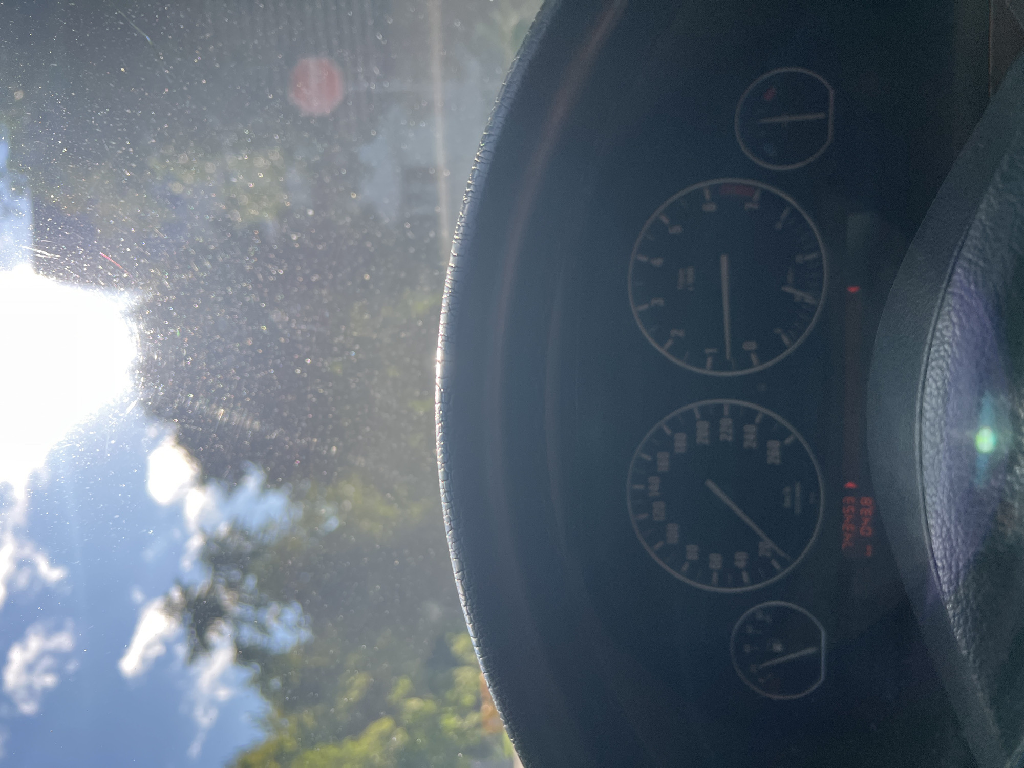 The image shows a view from inside a car, looking out through the windshield. The sun is bright and high in the sky, with some clouds present. Trees and buildings are visible in the background, but they are out of focus due to the glare and the view through the windshield. The steering wheel and dashboard are in the foreground, with the speedometer and tachometer visible. The car's odometer displays "243453" kilometers, and the trip meter shows "0438". The image appears to have been taken in a rural or suburban area.