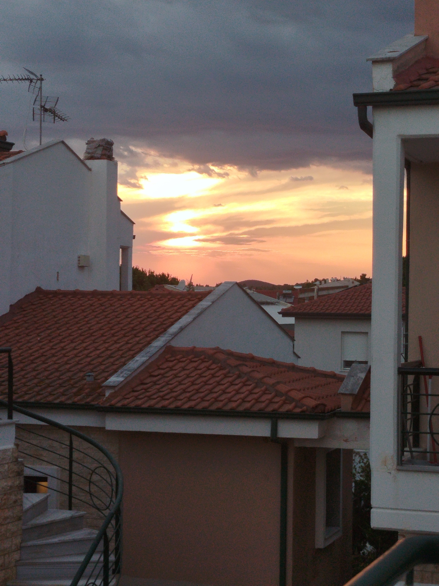 The image depicts a sunset scene over a residential area in Sithonia Municipal Unit, Greece. The sky is filled with dramatic clouds, with the sun breaking through to cast warm orange and yellow light across the horizon. The foreground is dominated by the rooftops of several houses, characterized by their reddish-brown tile coverings. The architecture suggests a Mediterranean style, with white-washed walls visible on some buildings. A spiral staircase with a dark metal railing is visible on the left, leading up towards one of the houses. In the background, a scattering of trees and other buildings can be seen against the colorful sky. There are no people or animals visible in the image. The scene is static and captures a moment of natural beauty at dusk.