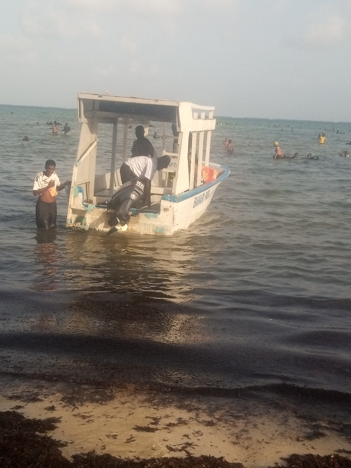 The image depicts a coastal scene in Shanzu ward, Kenya. In the foreground, a dark, wet shoreline covered with abundant dark seaweed transitions into calm, greenish-blue water. A small, white boat with a blue trim and a rectangular white canopy structure is positioned in the shallow water, facing away from the viewer. An outboard motor, dark gray with a silver propeller, is mounted at the boat's stern, partially submerged. Text "BARAKAH 2" is visible on the hull, along with "CAPT" or "PTI" near the canopy. An orange life preserver is inside the boat.
Three individuals are in and around the boat. A male, dark-skinned person wearing a white t-shirt and dark shorts is standing in the water to the left of the boat, holding a yellow object and a white cloth. Inside the boat, a second male, dark-skinned person in a white t-shirt and dark clothing is bent over at the stern, attending to the outboard motor. A third dark-skinned person is visible as a silhouette standing under the boat's canopy.
In the mid-ground and background, numerous other individuals are dispersed across the shallow water, appearing to swim or wade. Some are partially submerged, and a few wear yellow head coverings. The water extends to a hazy horizon under a bright, light blue sky.