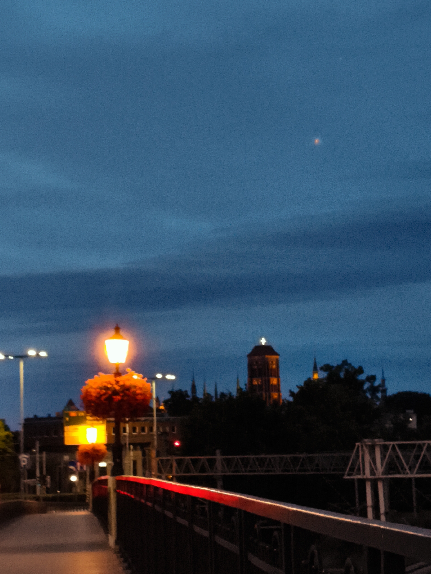 The image captures a twilight view of the Motława River waterfront in Gdańsk, Poland. In the foreground, a red-railed bridge spans the water. To the left, a lamppost with an illuminated orange light stands amidst red floral decorations. Further in the background, a prominent brick tower, likely part of the historic city architecture, is visible, illuminated by interior lights. The sky above is a deep blue, with a faint, distant light appearing in the upper right quadrant, potentially a planet or a distant light source. The overall scene depicts a tranquil evening cityscape.