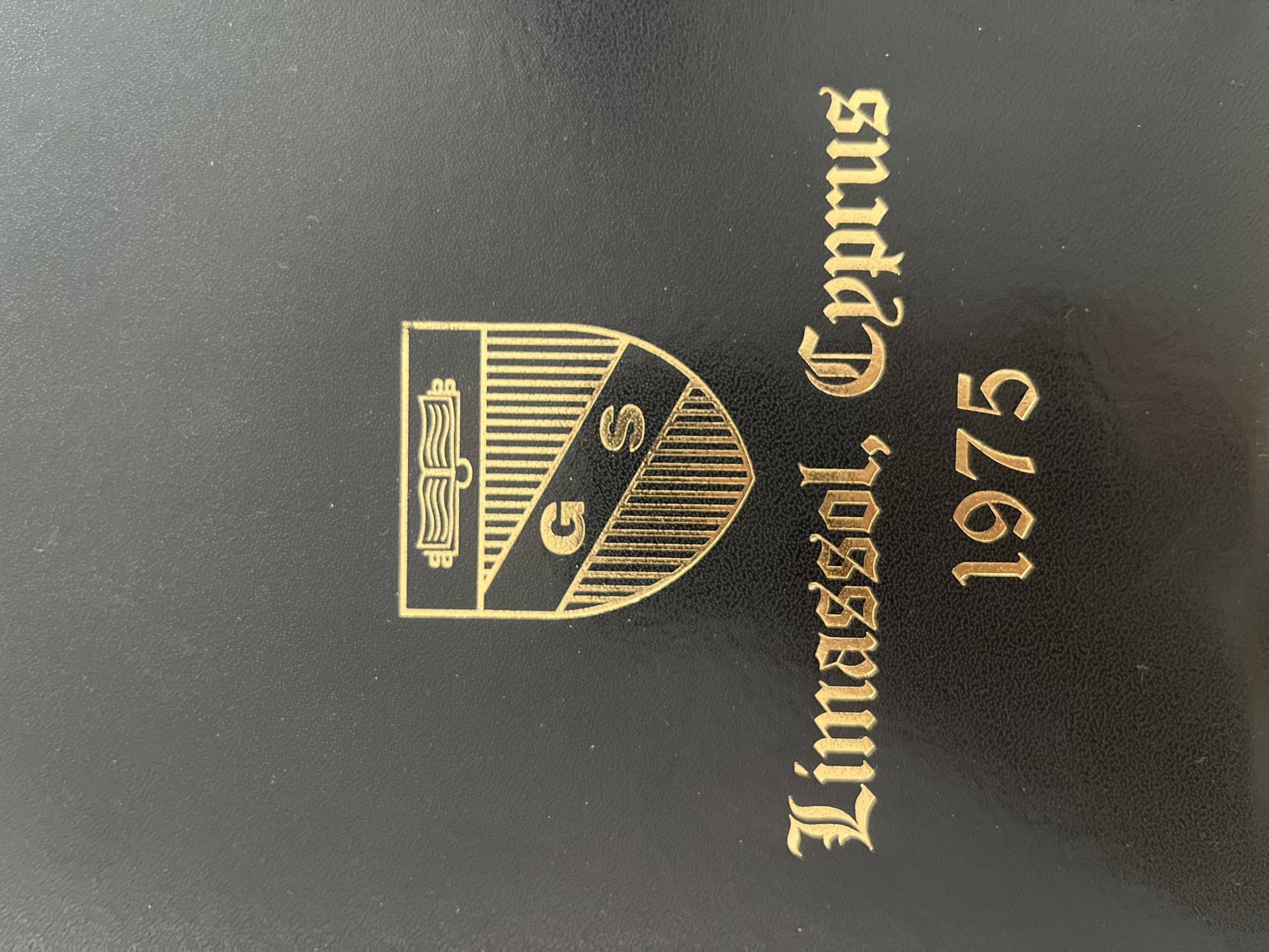 The image shows a black surface with gold lettering and a gold crest. The crest contains an open book at the top and a shield divided into sections with vertical lines. Inside the shield, there are the letters "G" and "S". Below the crest, the text "Limassol, Cyprus" is written in an ornate font, followed by the year "1975". The overall appearance suggests a commemorative plaque or cover. There are no people, actions, or interactions depicted. The environment is the surface itself, which appears to be textured. The location is identified as Limassol, Cyprus.