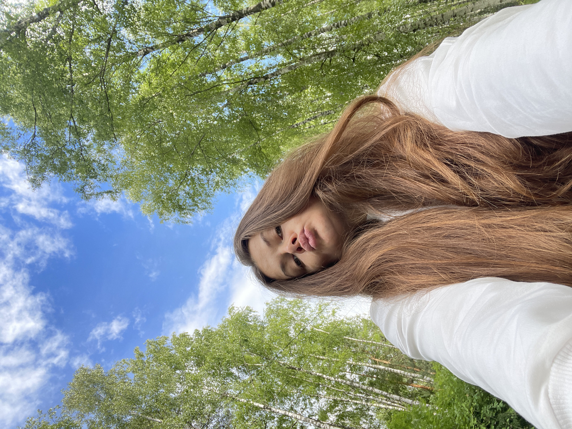 A young woman with long, brown hair is captured in an upside-down selfie taken outdoors. She is wearing a white long-sleeved shirt. Her face is turned towards the camera, with her lips slightly pursed. The background consists of a vibrant blue sky with scattered white clouds, and the lush green canopy of trees, likely birch trees given the location context of Заречный, Russia. The sunlight suggests it is daytime, possibly late morning or afternoon, with bright but not harsh lighting. The overall mood appears casual and relaxed, with the woman enjoying a moment outdoors amongst nature. No visible text is present in the image.
