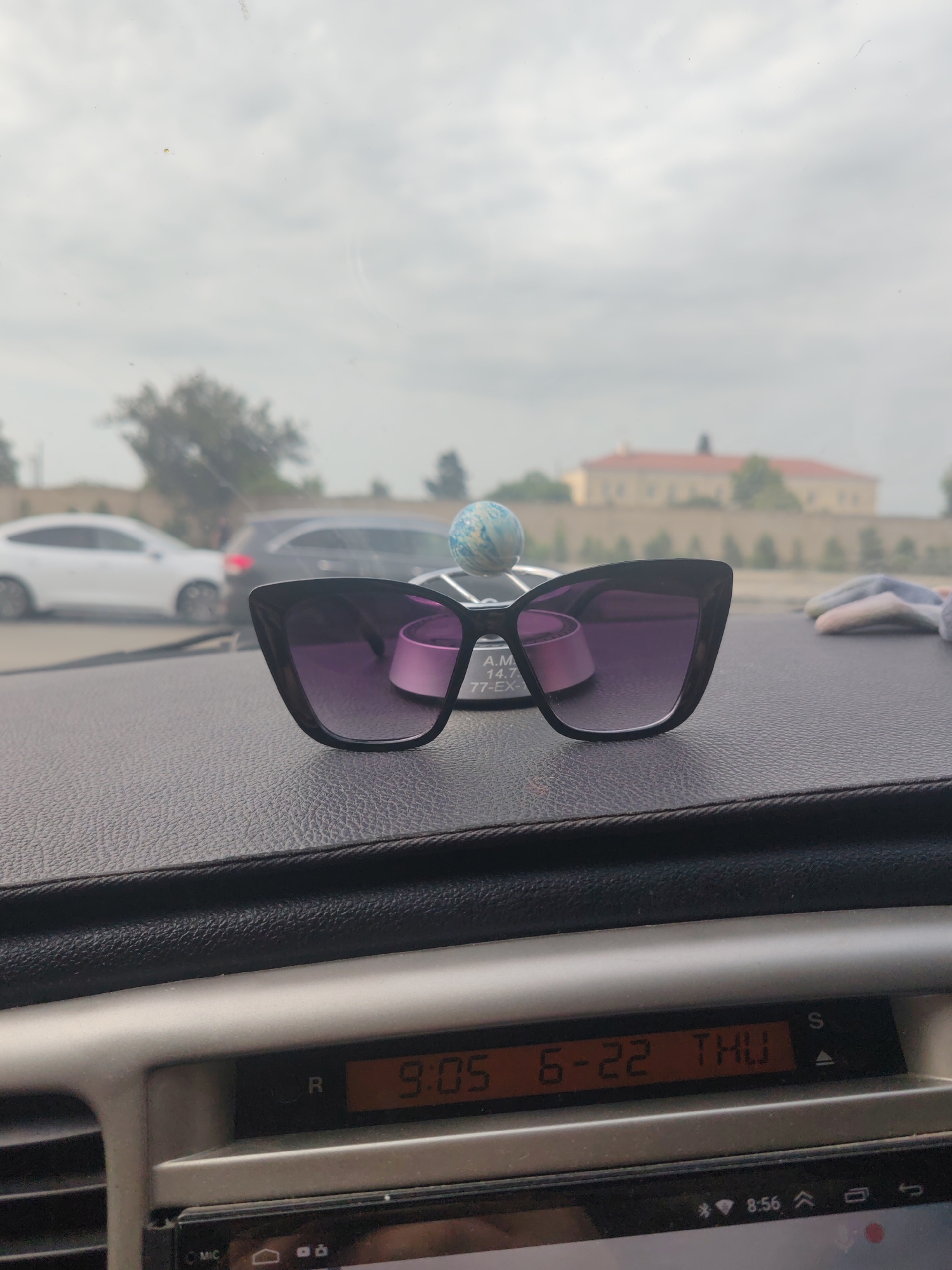 The image shows a pair of black cat-eye sunglasses with purple lenses resting on a dark, textured dashboard inside a car. A small, blue and white marbled globe ornament is positioned behind the sunglasses, and to the right, a grey and white patterned fabric item is visible. In the background, through the car's windshield, a street scene is blurred. Several cars are on the road, including a white sedan and a dark SUV. Beyond the road, a building with a red roof and light-colored walls is visible, surrounded by trees and a light-colored wall. The sky is overcast. On the dashboard, a digital display shows the time as "9:05" and the date as "6-22 THU". The car is likely in Baku City, Azerbaijan, given the presence of the car's dashboard display showing the time and date in a format common for the region.