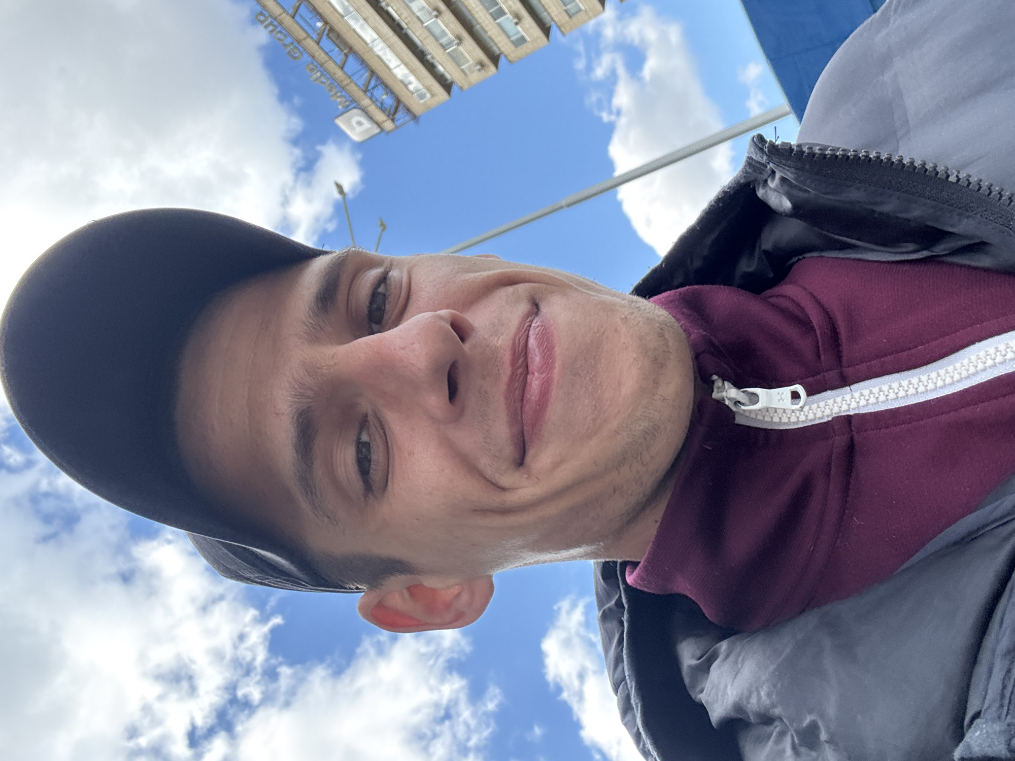 A man wearing a black cap, maroon hoodie, and dark gray jacket is taking a selfie. The sky above is blue with scattered clouds. A tall building is visible in the upper background. The building has a sign that reads "d media group." The photo was likely taken in Sofia, Bulgaria.
