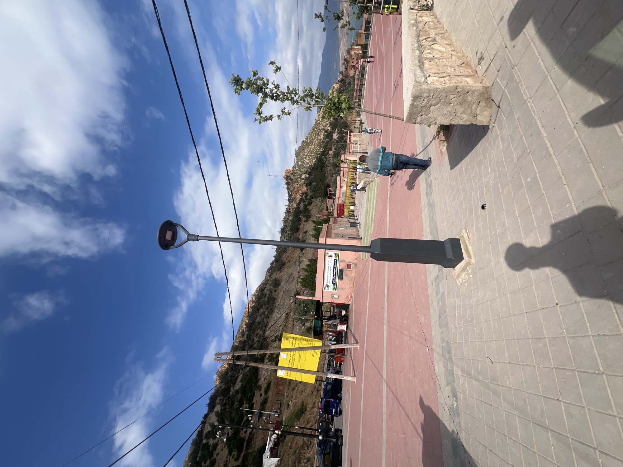 The image depicts a daytime scene in Bin El Ouidane, Morocco. A paved walkway transitions from red to gray tile. A black lamppost stands prominently on the gray tiled area. A mountain rises in the background, with buildings and trees visible along its slopes. A building with signage and posters stands adjacent to the red paved area. A yellow sign is propped up by wooden supports next to parked cars. Utility lines run across the sky. A person in a blue jacket walks in the middle ground. The sky is blue with scattered clouds. A shadow cast by a person taking the photo is visible in the lower right corner.