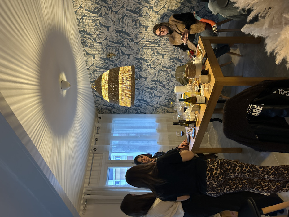 A casually set dining table displays wine bottles, glasses, and scattered items, suggesting a recent gathering. Three people are near the table, one in leopard-print pants and a black top, another in black, and a third in white. A woman sits at the table's far end, wearing glasses and a tan sweater. A distinctive lamp casts radiating shadows on the ceiling. Wallpaper with a blue leafy pattern adorns the wall, and natural light filters through a window.