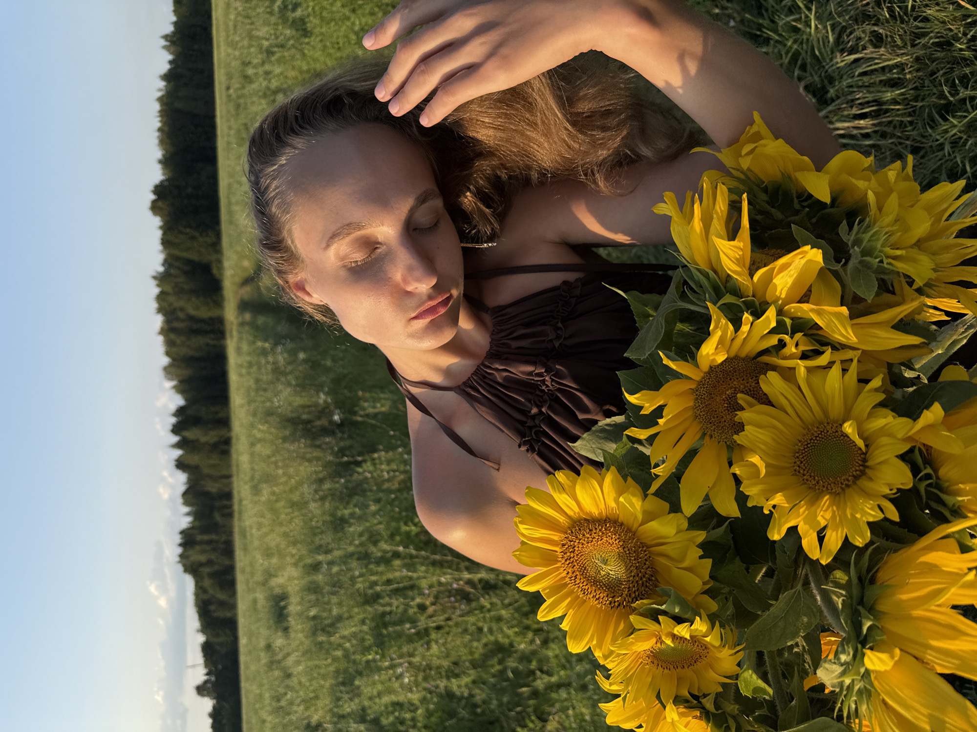 A young woman with blonde hair is in a field of green grass holding a bouquet of sunflowers. She is wearing a brown halter top and her eyes are closed as she tilts her head back. The sun is setting, casting a warm glow on the scene. In the background, there is a line of trees and a clear blue sky. The image appears to have been taken in Aksaitovo, Russia.