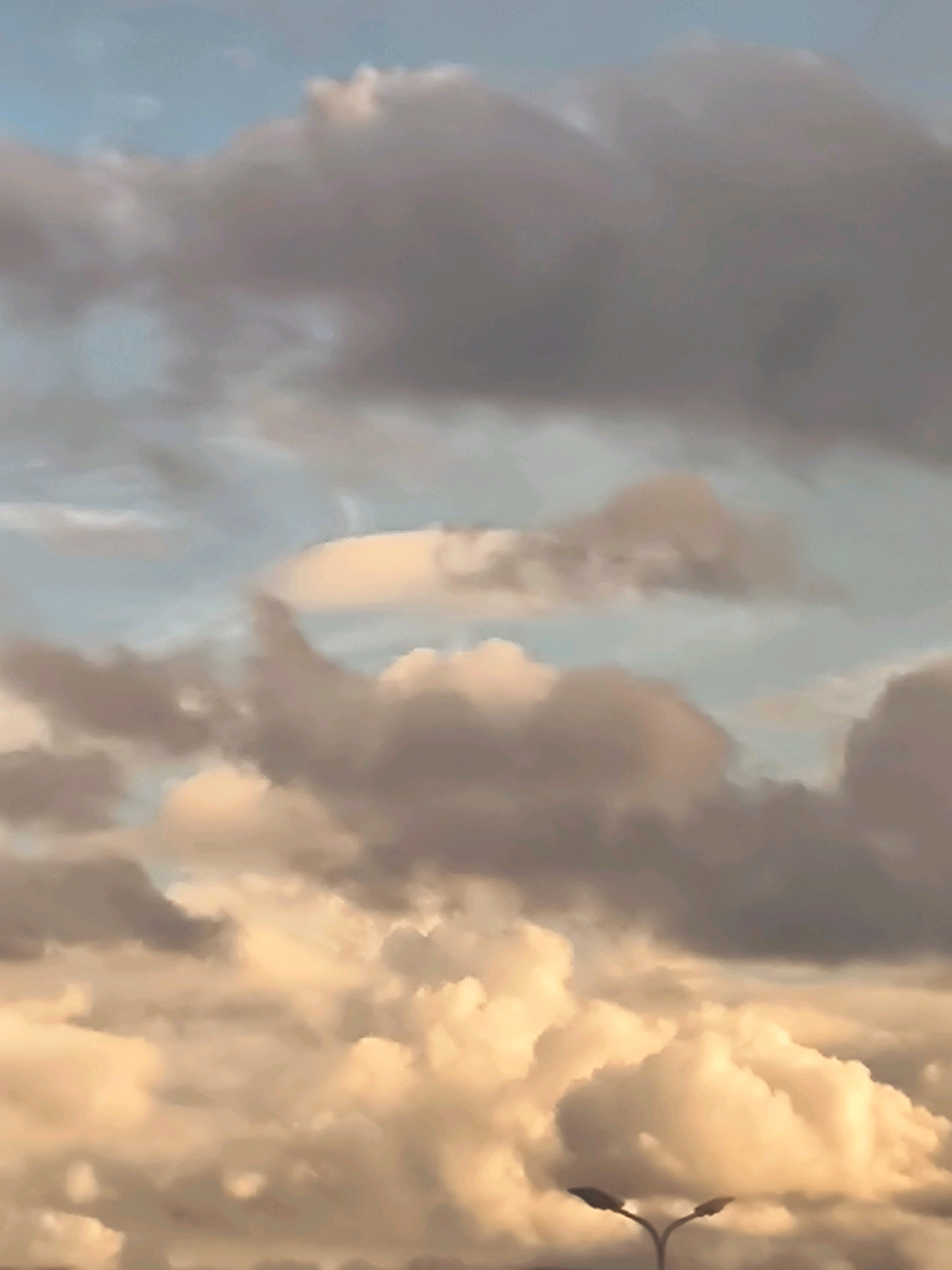 The image depicts a cloudy sky with a warm, diffused light, suggesting either sunrise or sunset. The clouds are a mix of soft grays and pale oranges, with some brighter, more luminous white areas that appear to be illuminated by the sun. The overall impression is one of a dramatic or atmospheric sky. In the lower portion of the image, two streetlights with a distinctive Y-shaped pole are visible against the clouds. The streetlights are dark and silhouetted. The scene appears to be an outdoor urban environment, possibly overlooking a road or a park, given the presence of streetlights. There are no people visible in the image. Without additional context or specific landmarks, it is impossible to pinpoint the exact location within Riga, Latvia.
