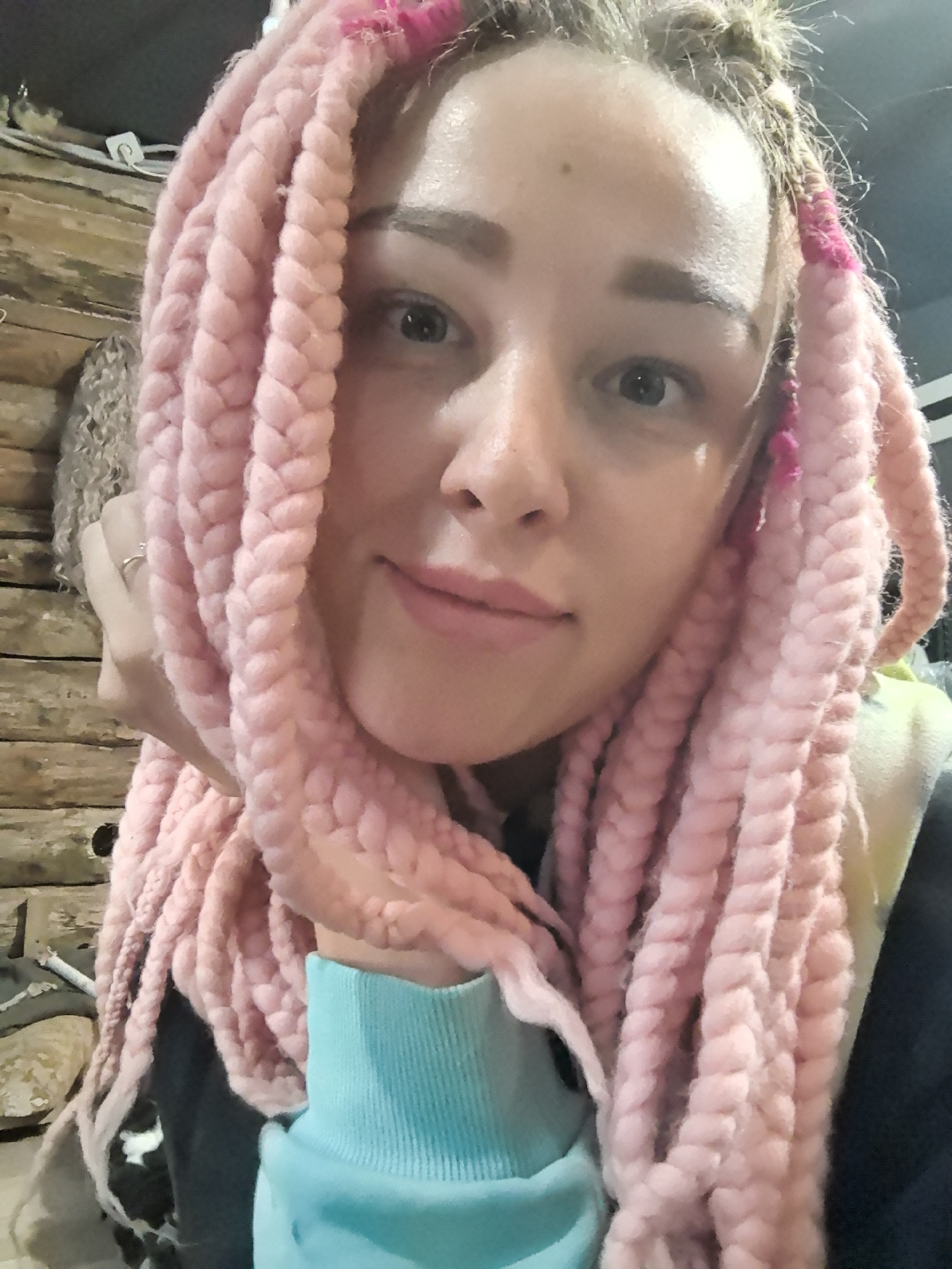 A close-up selfie depicts a fair-skinned individual with light eyebrows and eyes. The person has pink braids framing their face, with some strands adorned with pink accents. The person's left hand, sporting a ring, is gently supporting their head. They are wearing a garment with a light blue cuff.
The background reveals a log cabin interior, and a partially obscured person. The scene is indoors.