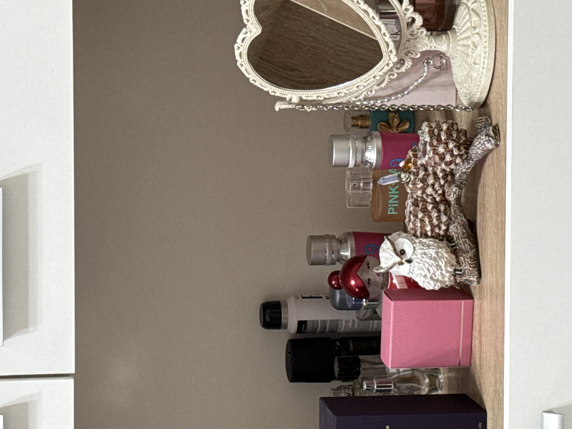The image shows the interior of a shelf or cabinet, filled with a collection of various items. At the top, there's a heart-shaped mirror with an ornate white frame. Below the mirror are several bottles of perfume or cosmetics, including a bottle with a pink label. An owl figurine, appears to be perched on a stack of items. Further down, there's a pink box and various other containers of different shapes and sizes, some with black caps or labels. At the very bottom, a dark blue box with gold lettering. The shelf's surface has a light wood grain appearance. The overall setting suggests a personal storage space within a home.