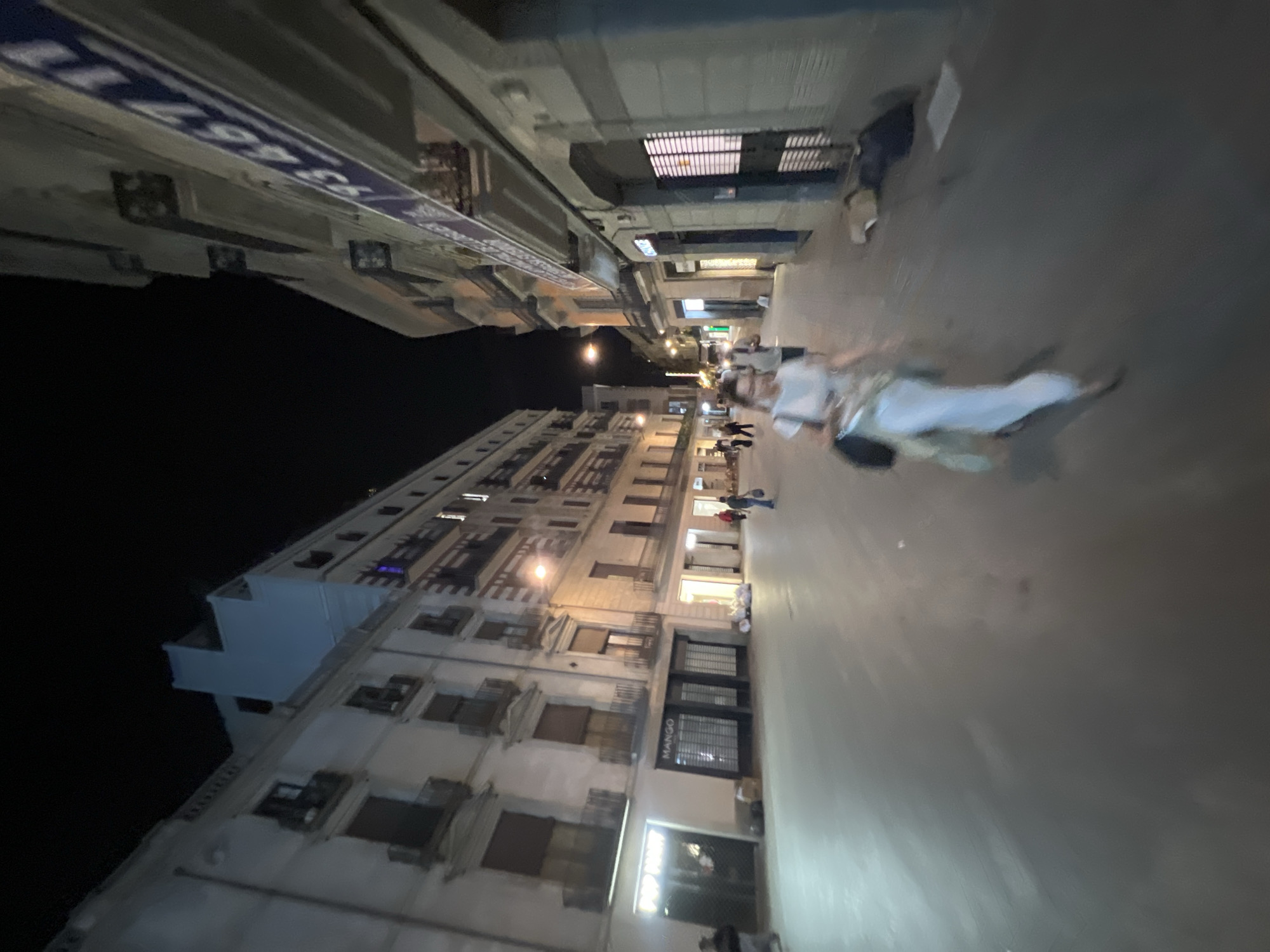 A nighttime street scene in Barcelona, Spain, captures a pedestrian-only thoroughfare lined with buildings. The street is paved, and the architecture suggests a European city. On the left, a white building with multiple balconies is illuminated. Shops are visible at street level, including one with a "MANGO" sign. A few individuals are present, walking in different directions. In the foreground, a woman in a light-colored dress is blurred due to motion. Further down the street, other figures and shopfronts are visible. The scene is lit by streetlights and the illumination from shop windows, creating a nocturnal urban ambiance.