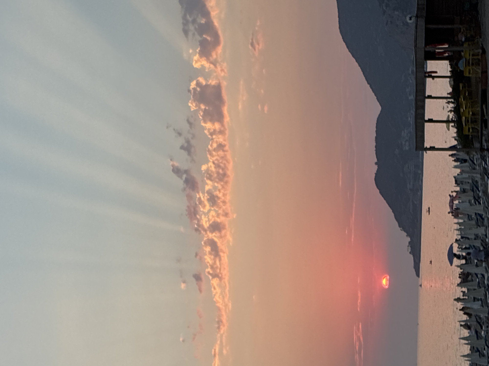 The image captures a sunset scene at a beach in Alanya, Turkey. The sun, a vibrant red orb, is partially submerged below the horizon, casting a warm glow across the sky. Sunbeams fan out from behind the sun, creating a dramatic effect. The sky transitions from a soft pink near the horizon to a pale blue at the top, with scattered clouds tinged with orange and pink. In the foreground, a beach is set up with numerous white beach chairs and umbrellas, some of which are open. A few people are visible sitting or standing on the beach, and several small boats are dotted on the calm sea. In the middle ground, a dark, silhouetted mountain range forms the backdrop. On the right side of the image, a rustic beachside structure with a wooden roof and yellow tables and chairs is partially visible, suggesting a cafe or bar. The overall atmosphere is tranquil and picturesque.