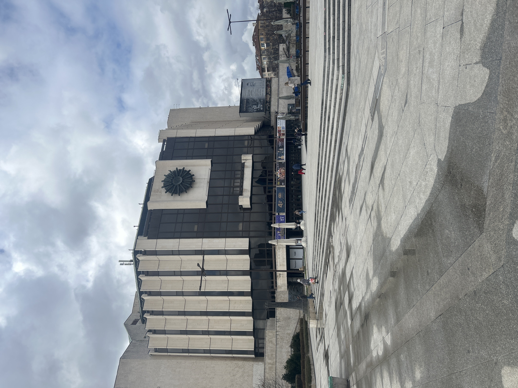 This image shows the National Palace of Culture in Sofia, Bulgaria. The building is a large, modern structure with a distinctive sun-shaped decoration on its facade. It is made of light-colored stone and glass. A wide, stepped plaza extends in front of the building, with people walking and standing on the plaza. The sky is cloudy and overcast. In the background, other buildings can be seen.