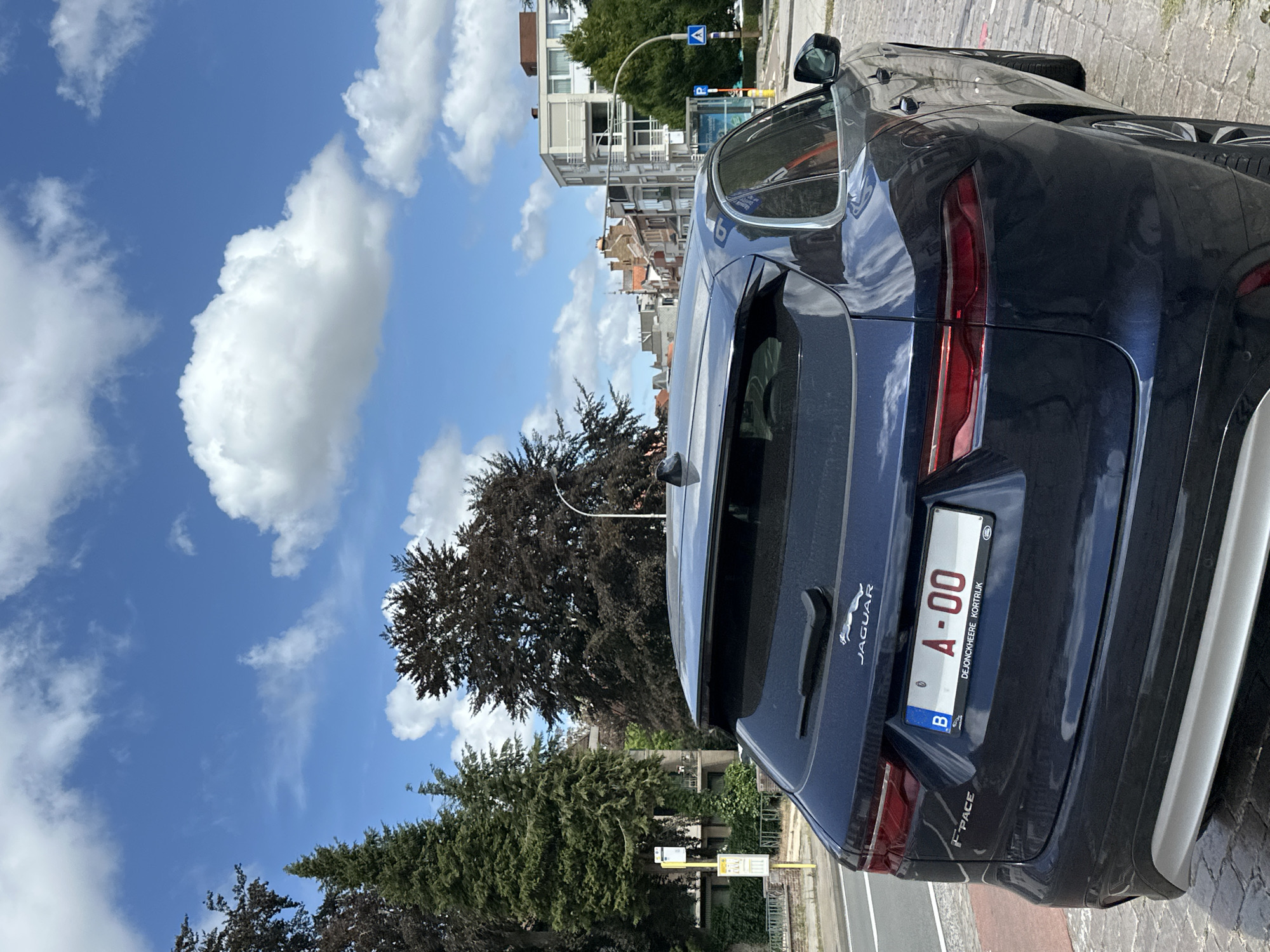 A dark blue Jaguar F-Pace is parked on a cobblestone street. The car has a Belgian license plate with the characters "A-00" and the text "DEJONCKHEERE KORTRIJK". The car is facing away from the camera. In the background, there are trees, buildings, a bus stop, and a street light under a bright blue sky with white clouds. The scene appears to be in a European city, possibly Bruges, Belgium.