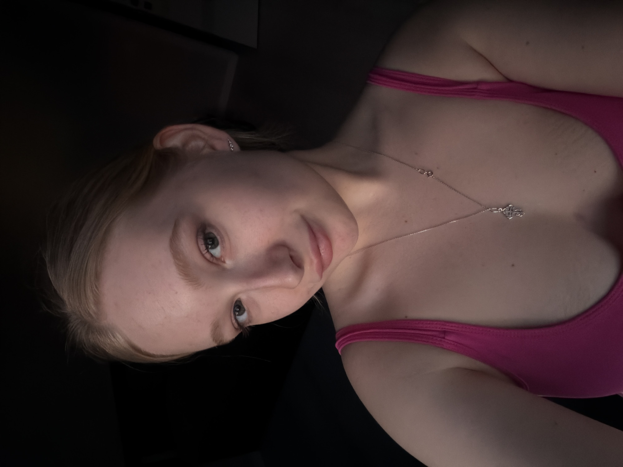 A fair-skinned woman with blond hair is pictured in a selfie. She is wearing a pink tank top and a silver necklace with a pendant. The background is dark, with a hint of what appears to be a cabinet in the upper left corner. The location is likely indoors. There is no definitive indication of Rossiyskiy, Russia.