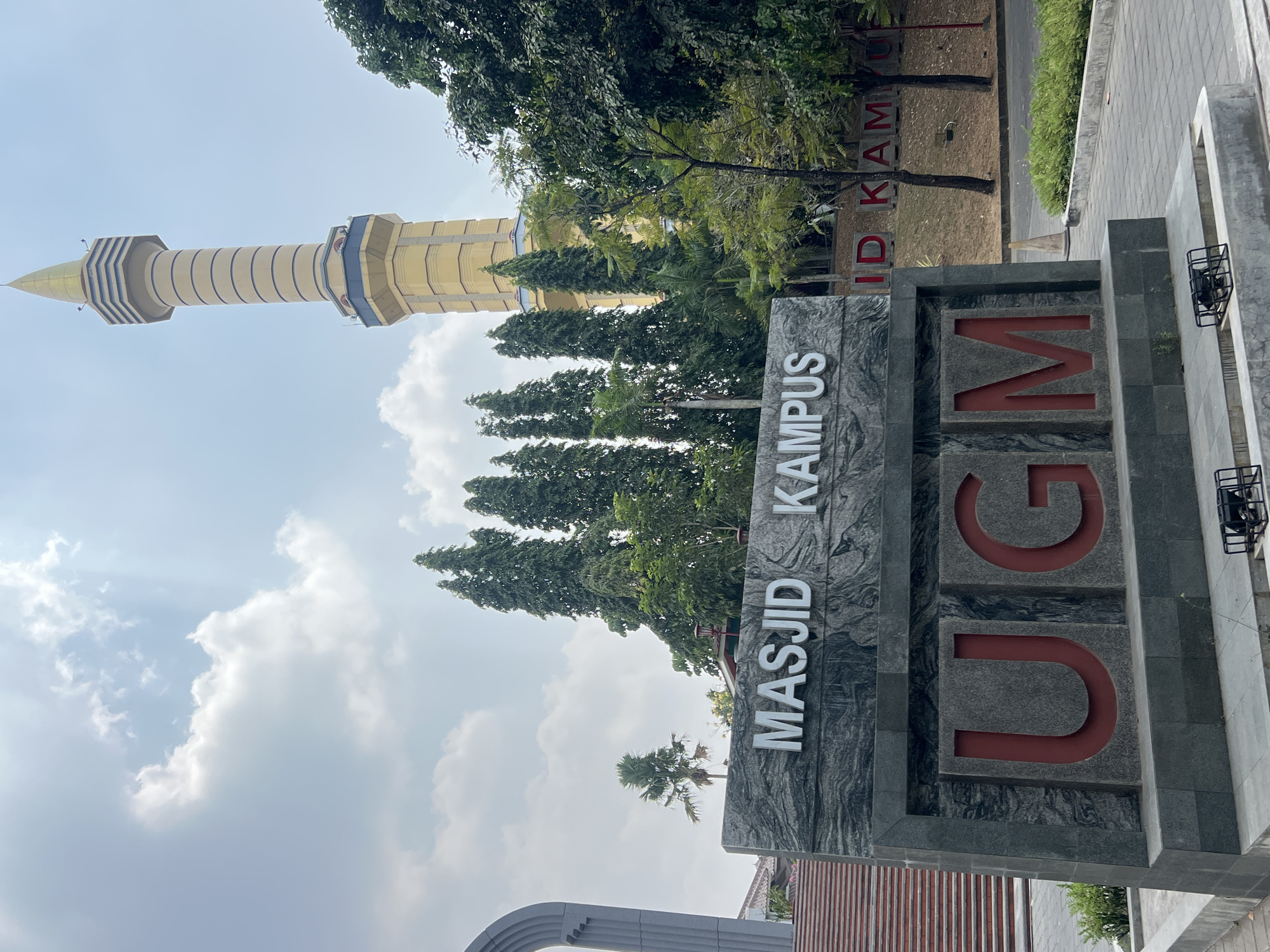 The image shows a monument sign with the text "MASJID KAMPUS UGM" in English, which translates to "Campus Mosque UGM". The monument is made of grey stone and has large red letters spelling out "UGM". Behind the monument, a tall, golden minaret of a mosque rises into a cloudy blue sky. Several tall green trees and palm trees are also visible. The scene appears to be an entrance or landmark on a campus. This is a photograph taken in Depok, Indonesia.