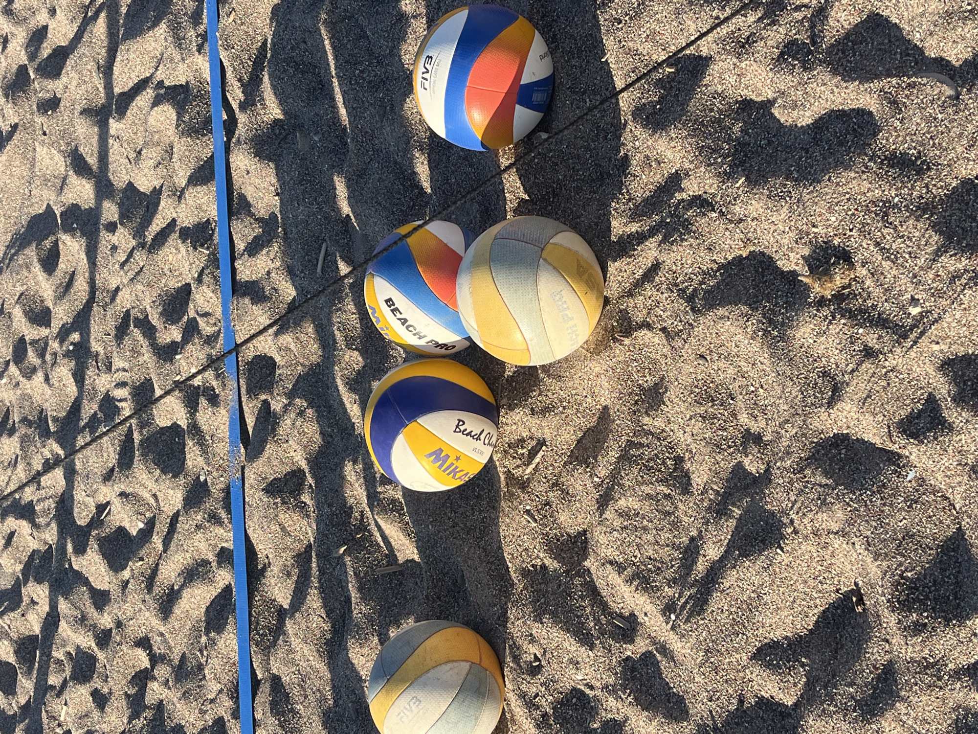 The image depicts a beach volleyball scene. Five volleyballs are scattered on a sandy surface, adjacent to a blue boundary line. The volleyballs vary in color and branding, with brands such as FIVB and Mikasa Beach Champ visible. The sand appears coarse and is partially shaded. The scene is outdoors. The location is the city of Budva, Montenegro.