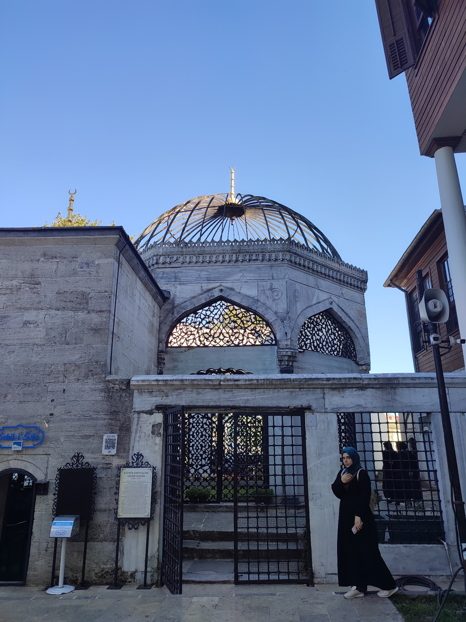 A woman in a black abaya and blue hijab stands in front of a historic building with a dome in Üsküdar, Turkey. The building features stone walls and intricate metalwork on its windows and gates. A sign with Turkish text is mounted on the wall. The dome is made of glass and metal, topped with a crescent moon. To the right of the building, a wooden structure with shutters is visible, and a speaker is mounted on a pole. The sky is clear and blue.