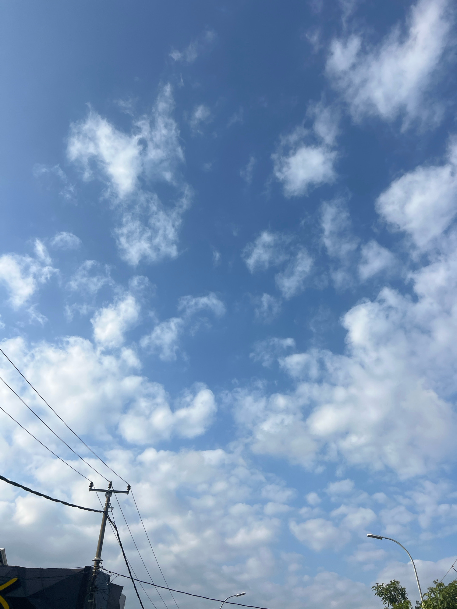 A low-angle, vertical shot captures a bright blue sky filled with scattered white, cumulus clouds. In the lower left quadrant, a dark, angular structure, possibly a building or awning, is visible. A utility pole with insulators and wires stands prominently on the left side, with several dark electrical wires crisscrossing the frame from left to right. In the lower right quadrant, a modern street lamp with a silver curved arm extends into the sky, and a small section of green trees can be seen. The scene depicts an outdoor urban environment in Kuta, Indonesia, on a clear day with a few clouds.