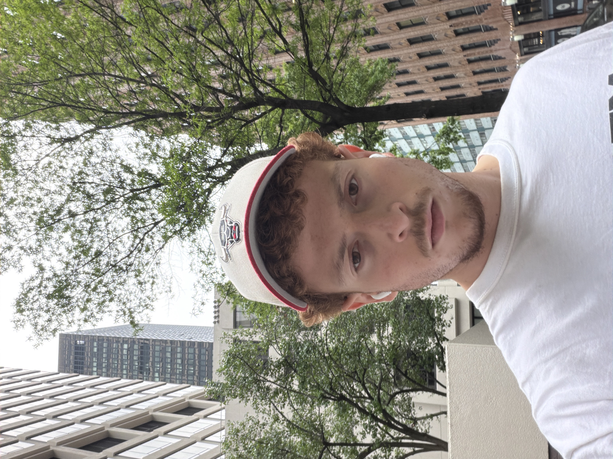 A close-up, slightly rotated view depicts a man wearing a baseball cap with a white crown and a red brim, a white t-shirt, and what appears to be a wireless earbud in his ear. He is positioned against a backdrop of urban architecture and greenery. The buildings in the background have a symmetrical window design with horizontal rows extending across the structures. Trees with green leaves are visible, partially obscuring the buildings. The man has curly reddish-brown hair and a beard with trimmed mustache. The setting appears to be an outdoor location in a city; due to architecture and the baseball cap of the Detroit Tigers, I will call it Detroit, United States of America.