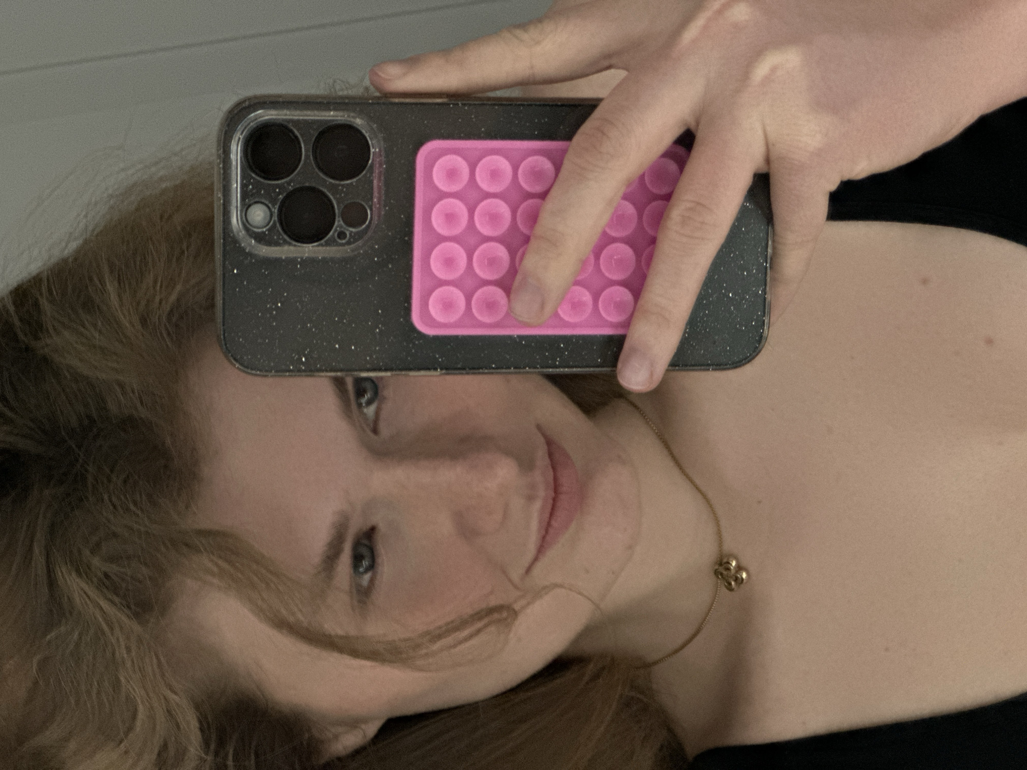 A person is taking a selfie. The person has light hair and blue eyes, and is wearing a gold necklace. The person is holding a black iPhone with a pink pop-it attached to the back. The location appears to be indoors. The city of Kazan, Russia, cannot be determined from the image.