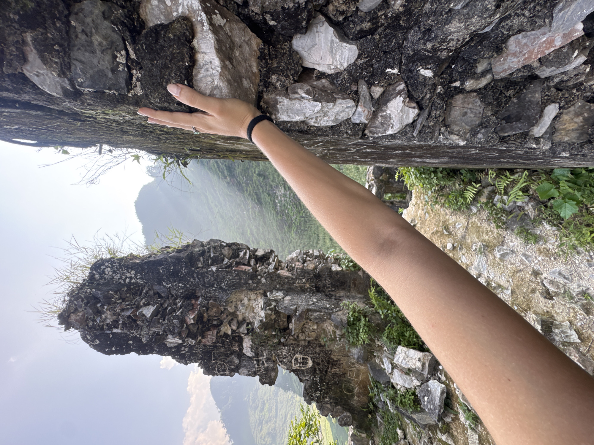 A medium shot depicts an arm, angled from lower right to upper left, reaching towards a rock wall composed of dark and light grey stones. The hand is positioned on the underside of the wall, fingers extended. A black hair tie is visible on the wrist. In the background, a landscape of mountains and green vegetation is visible. The location is Đường Thượng Commune, Vietnam.
