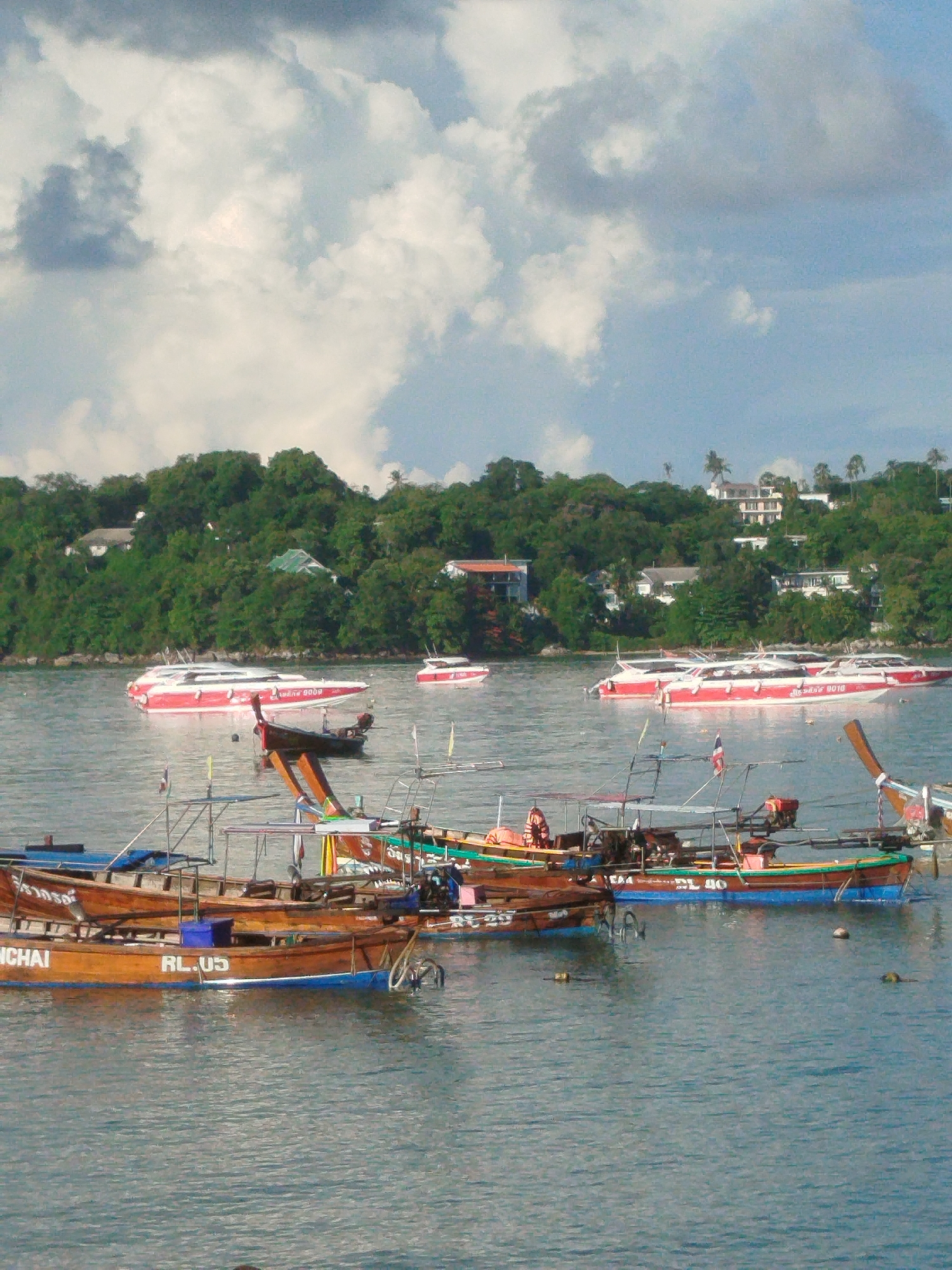 This is a vertical, eye-level, full-shot photograph capturing a busy harbor scene in Rawai, Thailand. In the foreground, several traditional Thai longtail boats, characterized by their wooden hulls and prominent outriggers with engines, are moored. One boat has the designation "RL.05" visible on its side. A person wearing an orange life vest is seated in one of the boats. In the mid-ground, several sleek, modern speedboats with red and white hulls are anchored or moving. These speedboats appear to be tourist vessels, some displaying Thai script. The background features a lush, green, tree-covered hillside dotted with houses and buildings, suggesting a coastal town. The sky above is partly cloudy with patches of blue sky visible. The water is a calm, muted blue-green, reflecting the sky and surrounding environment. The overall atmosphere is one of a vibrant maritime activity hub.
