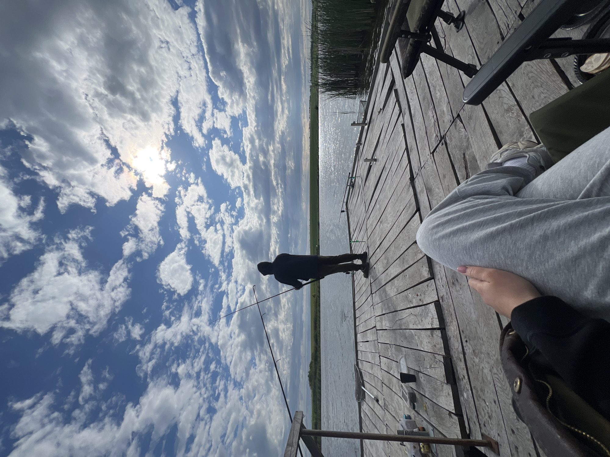 The image depicts a person sitting on a wooden dock, possibly fishing. Another individual stands further out on the dock, holding a fishing rod. The location is identified by the presence of a body of water and greenery in the background, consistent with a natural setting. The sky is filled with clouds, with the sun visible through them. The person sitting appears to be wearing gray clothing and white sneakers, while the standing individual is silhouetted. Other fishing gear and equipment are scattered on the dock.