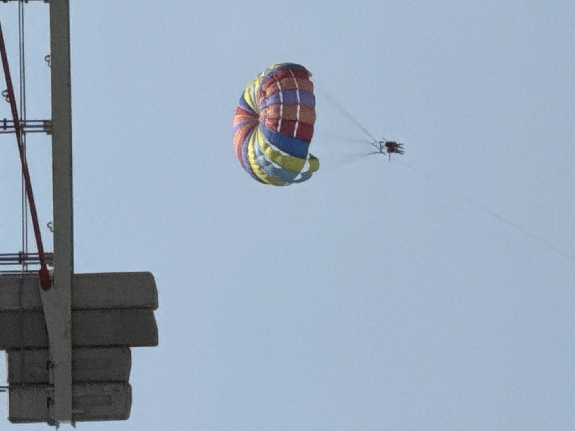 In Boreti, Montenegro, a parasailer is visible against a pale blue sky. The parasail is multi-colored with stripes of red, orange, yellow, green, blue, and purple. Attached to the parasail, two individuals are suspended in the air, likely enjoying a recreational activity. In the upper left quadrant of the image, a portion of a large, gray, concrete structure is visible, which appears to be part of a bridge or overpass. The structure has a horizontal beam and vertical supports.