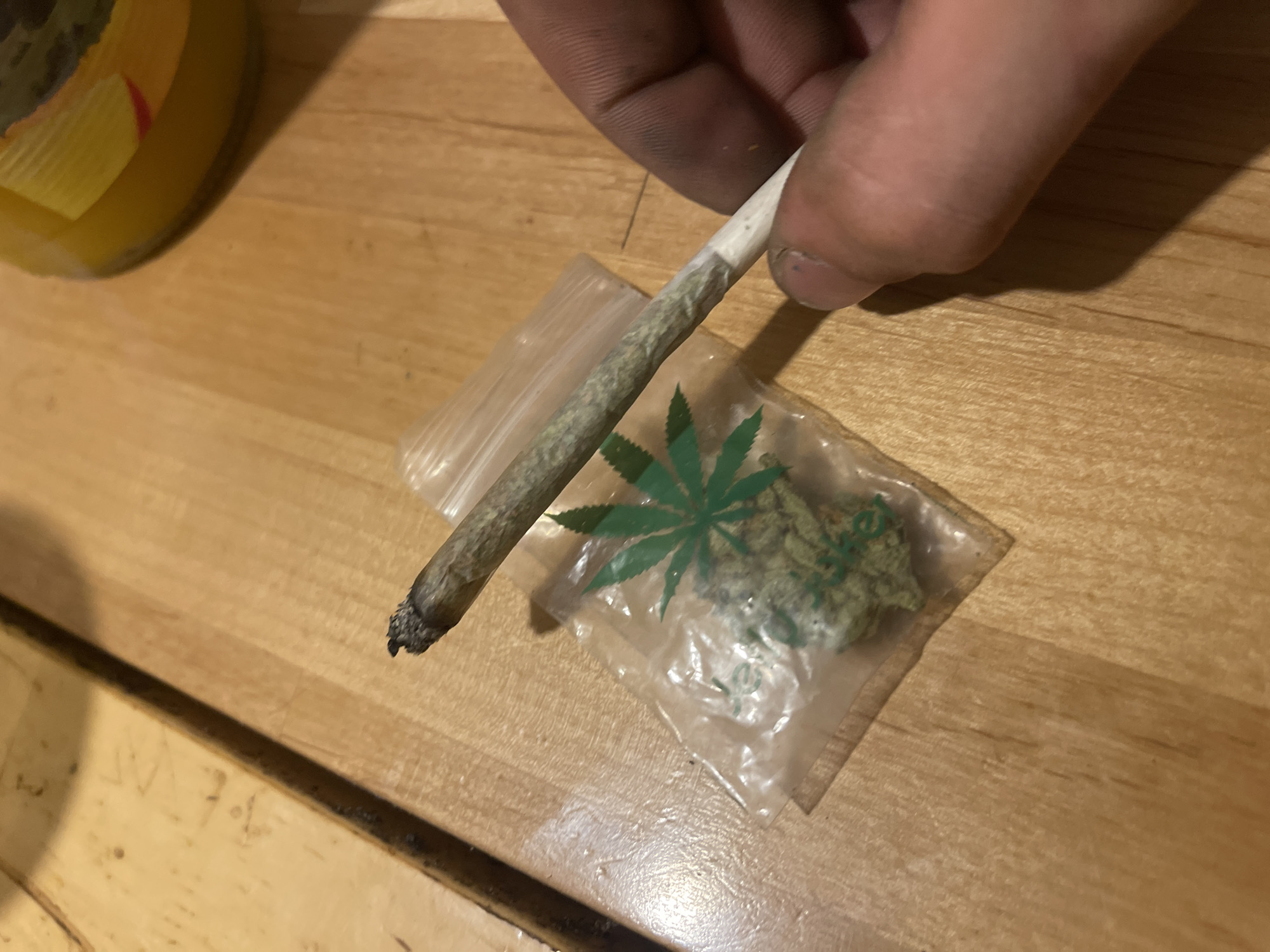 A hand is holding a lit marijuana cigarette on a wooden surface. Next to the cigarette is a small plastic bag containing marijuana buds. The background shows a blurry yellow object, possibly a jar. The scene appears to be indoors.
