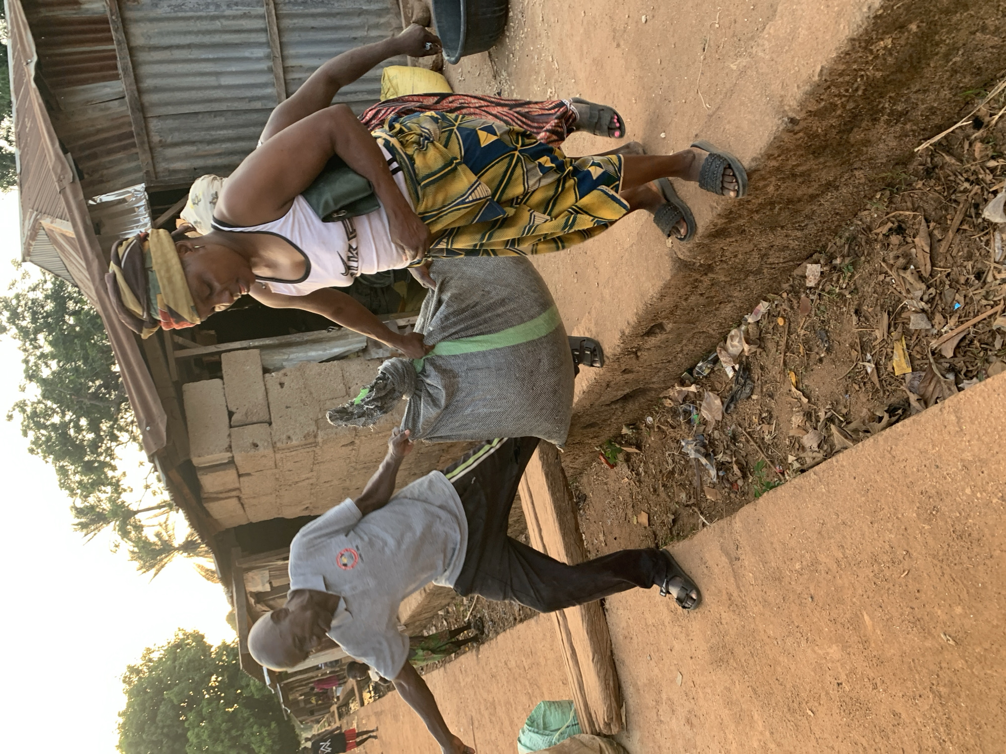 In Agban, Nigeria, two individuals interact outdoors. A man in a light grey t-shirt and dark pants extends his arm towards a large grey bag held by a woman. The woman, wearing a white tank top, a patterned yellow and blue skirt, and a head wrap, appears to be handing over the bag. The setting includes a building with a corrugated metal roof and a partially visible brick wall. The ground is a mix of dirt, debris, and a concrete path. Nearby, there are trees.