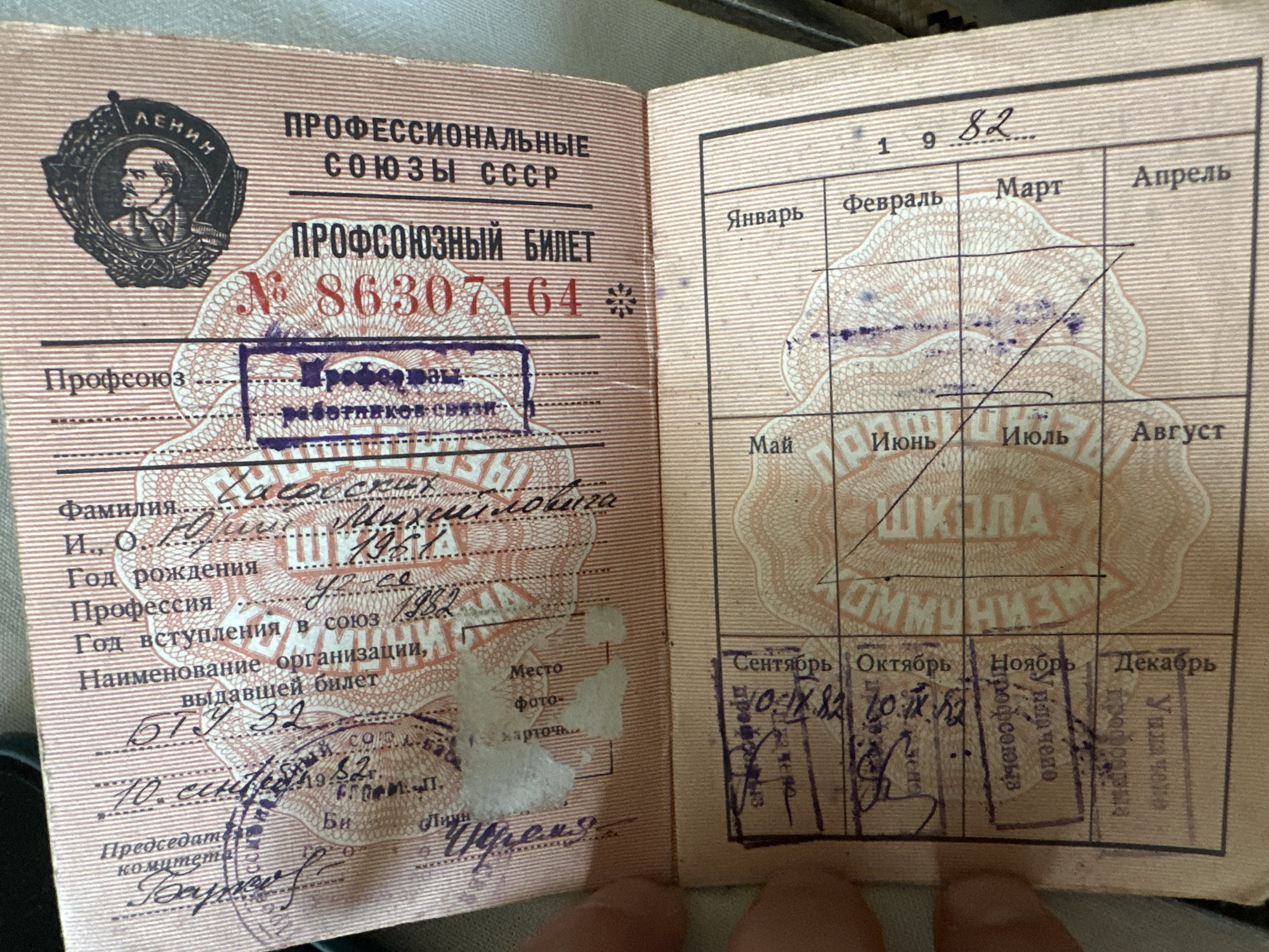 The image displays an opened Soviet-era trade union membership card. The card is pink with vertical red stripes. The left page contains a section for personal information including "Фамилия" (Surname), "И. О." (Initials), "Год рождения" (Year of birth), "Профессия" (Profession), and "Наименование организации, выдавшей билет" (Name of the organization that issued the ticket). There is a place for a photograph, labeled "Фото" (Photo), with some smudges and a faded stamp over it. There are also handwritten entries and stamps.
The right page is titled "ПРОФСОЮЗНЫЕ БИЛЕТ № 86307164 ПРОФЕССИОНАЛЬНЫЕ СОЮЗЫ СССР". Below this, there is a grid with months of the year, presumably for recording dues payments. The year "1982" is visible, with a handwritten entry "SX" following it, indicating the year the dues were paid. Several months have handwritten entries and stamps, some of which appear to be from the city of Biysk, Russia. Specifically, a stamp on the right page reads "Бийск" (Biysk). The stamps are purple and circular, with text that is mostly illegible due to ink bleed and overlap. One stamp on the left page appears to be a signature.
The scene appears to be an indoor setting with soft lighting, and a person's hand is visible in the lower left corner, holding the card open. The card is laid flat on a surface, and the focus is on the card itself.