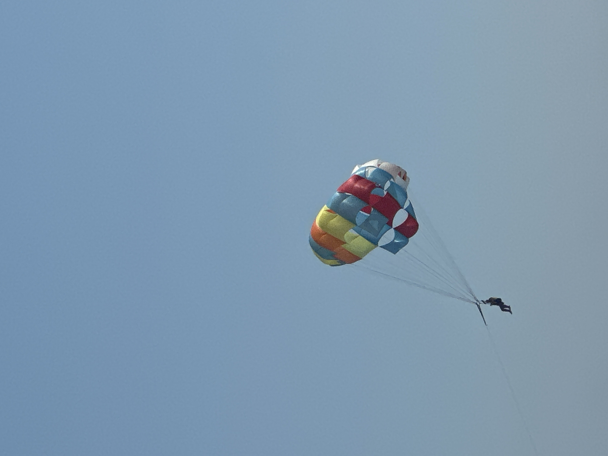 A lone individual is parasailing high in a clear, pale blue sky. The parasail, a large parachute-like canopy, is adorned with a vibrant pattern of colorful panels in red, blue, yellow, and orange, suggesting a festive and recreational activity. The person, dressed in dark clothing, is suspended beneath the canopy by a harness and lines, their posture indicating they are actively engaged in the sport. The background is a vast expanse of sky, devoid of any landmarks or specific geographical features that would pinpoint the location to Boreti, Montenegro. The overall scene evokes a sense of freedom and adventure, with the bright colors of the parasail providing a focal point against the monochromatic sky.