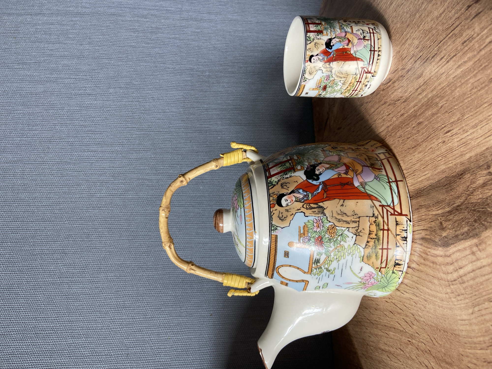The image displays a decorative porcelain teapot and a matching cup, both adorned with traditional East Asian imagery featuring figures in ornate clothing amidst a landscape of rocks, trees, and flowers. The teapot has a curved bamboo handle secured by yellow ties. The set rests on a wooden surface, likely a table, in front of a textured, blue wall. The scene is lit from above, casting subtle shadows. There is no indication of the city of Tuymazy, Russia, within the image.