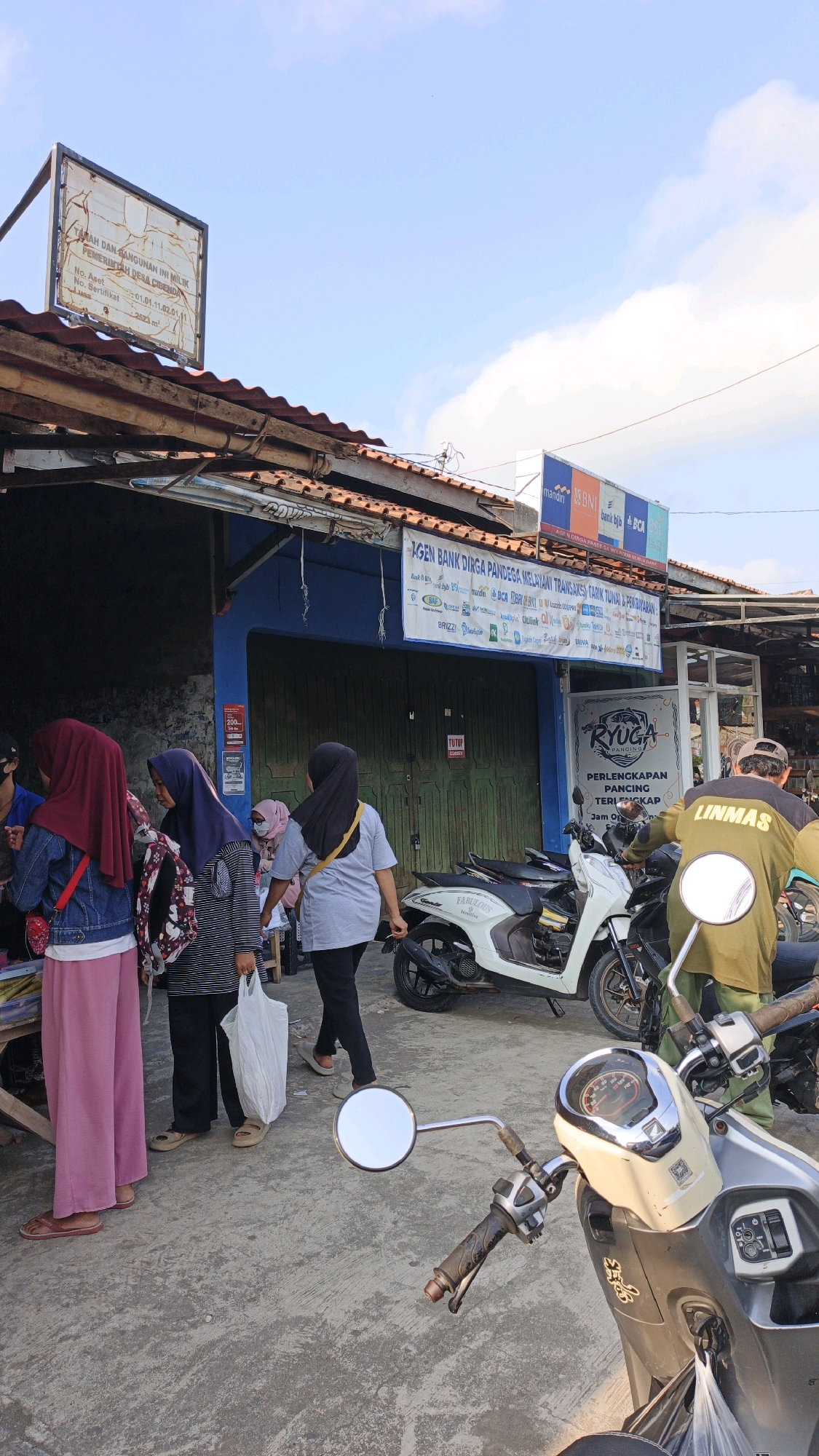 In Cibenda, Indonesia, an outdoor scene captures the facade of a small business with a blue-painted entrance and green metal doors. Above the entrance, a white banner displays "AGEN BANK DIRGA PANDEGA MELAYANI TRANSAKSI TARIK TUNAI & PERBANKAN" along with numerous logos of financial institutions. To the left of the entrance, a weathered white sign mounted on a wooden frame reads "TANAH DAN BANGUNAN INI MILIK PEMERINTAH DESA CIBEDA" followed by property details.
Several individuals are present. On the left, three women are gathered, one wearing a maroon hijab and denim jacket, another a patterned hijab and striped shirt, and the third a pale pink hijab and grey shirt. The woman in the patterned hijab is holding a white plastic bag. To the right of the entrance, a man wearing a green jacket with "LINMAS" printed on the back is seated on a motorcycle. In front of the business, two scooters are parked, a white one with "FABULOUS" written on its side and a darker scooter behind it. The ground is paved with concrete. The sky is partly cloudy with visible cumulus clouds. The overall impression is a busy commercial area on a bright day.