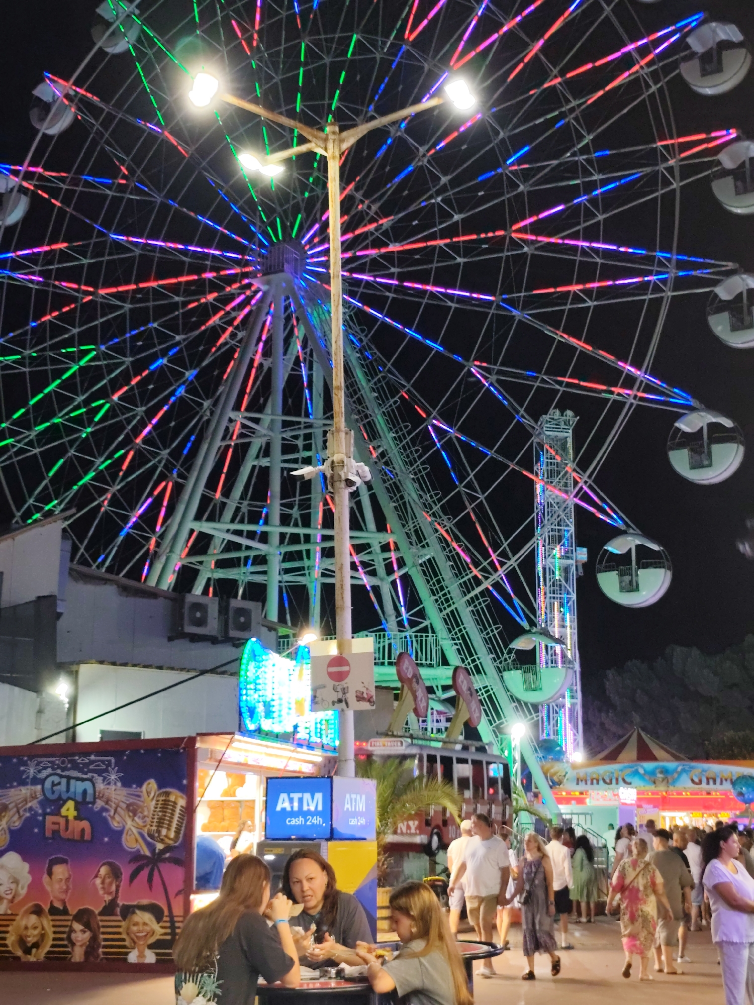 The image depicts an amusement park at night in Nesebar, Bulgaria. A large Ferris wheel dominates the scene, illuminated with colorful LED lights in red, green, and blue. In the foreground, a food stall with a "Gum 4 Fun" advertisement is visible, featuring cartoonish depictions of people and a microphone. Two women are seated at a table in front of the stall, engaged in conversation. To the right of the stall are two ATM machines, labeled "ATM cash 24h". A crowd of people are walking through the park, some in casual attire, suggesting a lively evening atmosphere. In the background, other amusement park attractions, including a tent with "MAGIC GAMES" signage, are discernible. The overall environment is characteristic of a summer fairground, with the darkness of the night contrasted by the bright lights of the attractions.