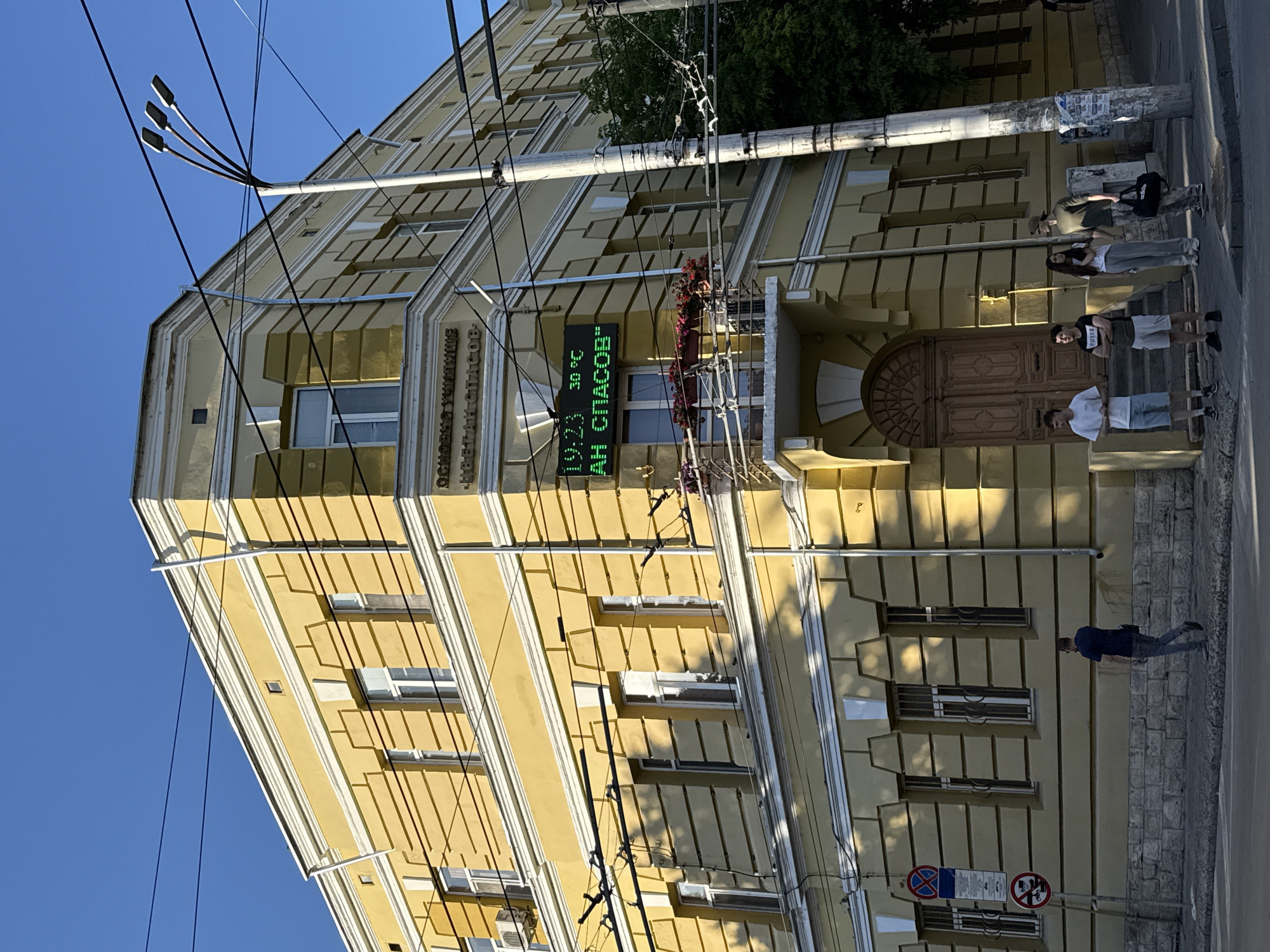 The image captures a corner view of a historic yellow building in Pleven, Bulgaria, bathed in the warm light of a late afternoon sun. The building features ornate architectural details, including prominent cornices and decorative window frames. A digital clock and thermometer display the time as 19:23 and the temperature as 30°C, with green LED characters spelling out "АН СПАСОВ" below. This likely indicates a business or institution named "AN SPASOV."
The building has several windows, some with visible window panes and air conditioning units. A balcony adorned with potted flowers extends from the upper floor. In front of the building, a prominent wooden double door serves as the main entrance, flanked by steps. A series of electrical wires crisscross the scene, suspended from a utility pole.
On the sidewalk, a group of five young individuals are gathered. From left to right, one young man stands facing forward, while another stands with his arms crossed. A young woman with long dark hair stands next to him, followed by another young man wearing a cap. The last individual in the group is partially obscured but appears to be carrying a black bag. They are casually dressed, suggesting a relaxed social gathering.
Further down the sidewalk, a single man in a dark blue t-shirt and jeans walks away from the camera. To the left of the building, a traffic sign with multiple panels is visible. The street in the foreground is paved, and a pedestrian crossing is indicated by white lines. The sky is clear and blue, typical of a summer day. The overall scene depicts a moment of everyday life in the city, with a blend of historical architecture and modern elements.