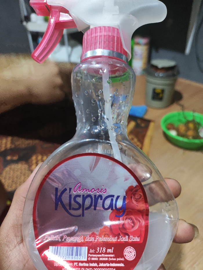 The image captures a close-up view of a clear plastic spray bottle, held by a person's hand, in an indoor setting in Pubian, Indonesia. The bottle, which is about half-full with a translucent liquid, features a pink and white spray nozzle with a transparent trigger. The person's thumb is visible on the left side, and fingers are visible gripping the bottle on the right.

The bottle has a distinctive red and white label with decorative elements. Prominently displayed is the brand name "Amoris Kispray" in purple and blue script, accompanied by an illustration of red roses. Below the brand name, the text "Pelícín, Pewangí, dan Pelembut Jadi Satu" translates to "Fabric Smoother, Fragrance, and Softener All in One," describing the product's function. The volume is clearly stated as "Isi: 318 ml."

Additional text on the label provides contact information: "Pertanyaan/Komentar: 08001 282828 (bebas pulsa)," indicating a toll-free number for questions or comments. The manufacturer is identified as "Diproduksi oleh PT. Herlina Indah, Jakarta-Indonesia," with a registration number "KEMENKES RI PKD 20203010734." A barcode is also partially visible on the label.

The background is blurred, suggesting a shallow depth of field, and shows various out-of-focus household items, including a patterned surface that might be a table or mat, a green bowl containing small, round objects, and other containers. The lighting indicates an indoor environment, likely under artificial light, as the time of day is not apparent. The overall scene suggests a casual, domestic setting.