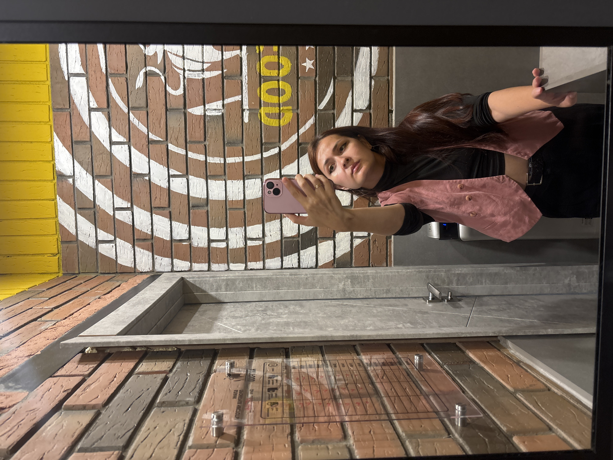 A young woman with long, dark hair takes a selfie in a restroom. She is wearing a black turtleneck, a pink vest, and black pants. She is holding a pink smartphone in her right hand. The restroom has a brick wall with a white mural, yellow ceiling accents, and a gray door. A clear acrylic chart is attached to the brick wall to the left of the door. The image appears to be taken in Naberezhnye Chelny, Russia, as suggested by the Russian text on the acrylic chart.