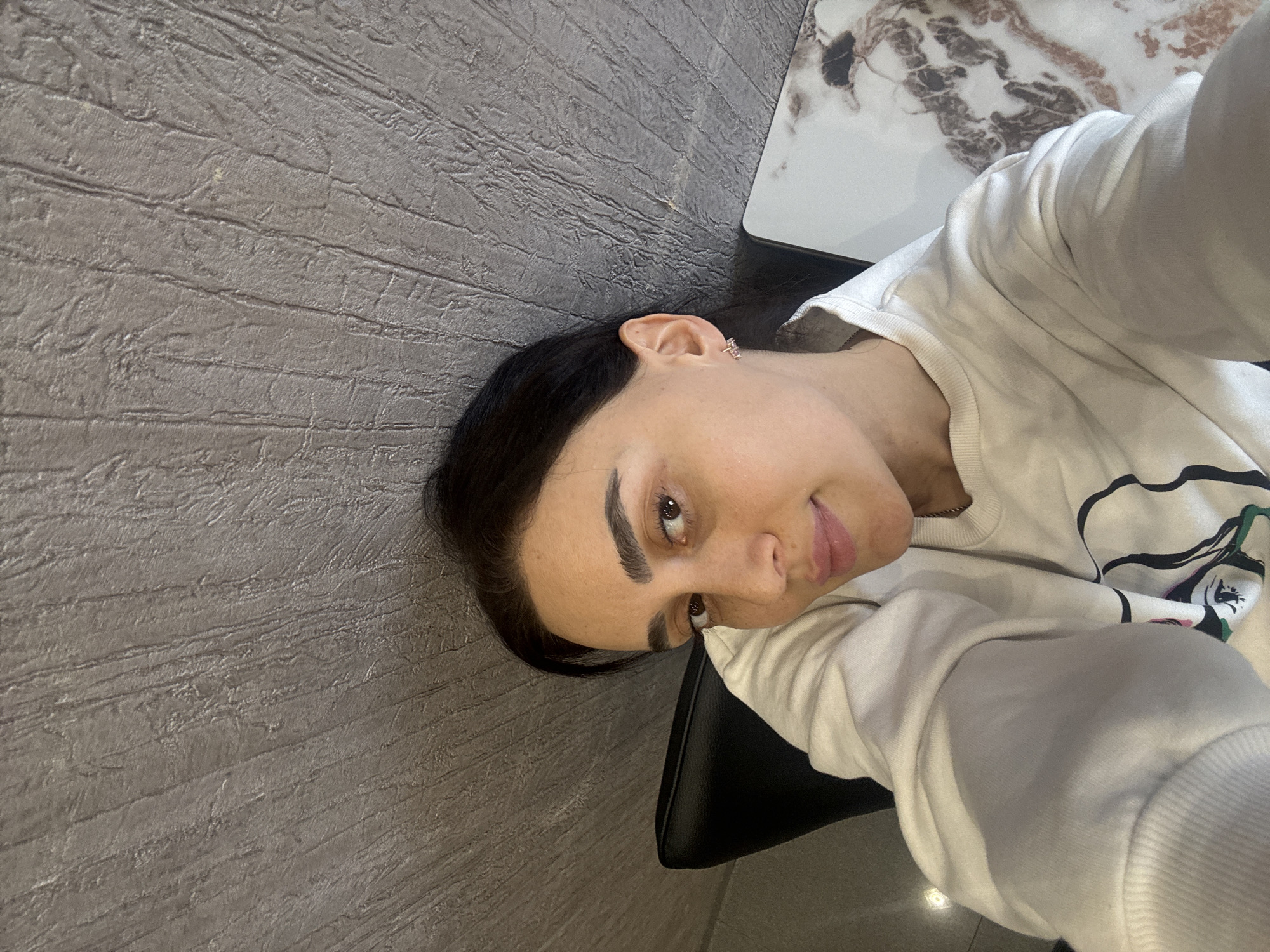 This media file captures a selfie of a young woman indoors in Tbilisi, Georgia. The woman, appearing to be in her 20s or early 30s, has dark hair, dark, well-defined eyebrows, and fair skin. She is looking directly at the camera with a subtle, neutral expression or slight smile. She wears a small, sparkly earring in her right ear. Her attire is a light-colored, long-sleeved top, possibly a sweatshirt, which features an abstract design with visible elements in black, green, and pink on the left side of her chest.
The background predominantly features a textured gray surface resembling a wall or floor with a grooved, wood-like pattern. To the right of the frame, a white and brown marble-patterned surface, likely a table, is visible. A black chair back or seat can also be seen behind the woman. The image has a distinct tilted angle, suggesting it was taken as a handheld selfie. The lighting indicates an indoor setting, likely artificial. There is no discernible text within the image. The overall impression is casual and composed.
