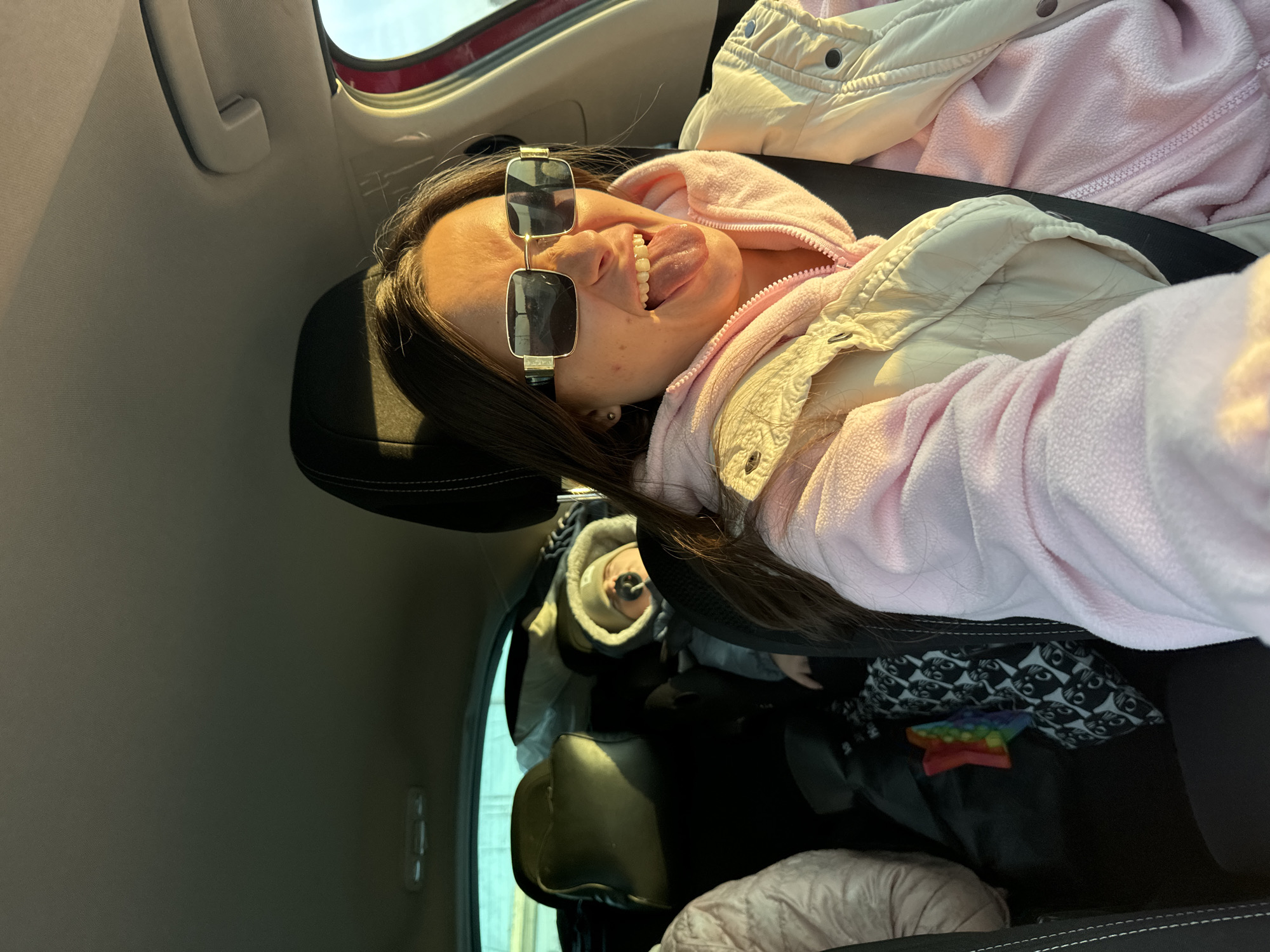 Inside a moving vehicle in Moscow, Russia, a young adult female is prominently featured in the front passenger seat, oriented towards the camera. She has long dark hair, wears rectangular gold-framed sunglasses, a light pink fleece hooded jacket, and a light beige quilted vest over it. Her seatbelt is correctly fastened across her chest and lap. Her facial expression is playful, with her tongue extended. Sunlight from the passenger side window illuminates her face and upper body.
In the back seat, behind the driver, a child is visible, partially obscured, wearing a grey hooded jacket and a hat, with a pacifier in their mouth. The child appears to be resting or sleeping. Further left in the back seat, only a portion of a third person's light-colored clothing is visible. The rear passenger area contains luggage, including a black and white patterned bag and a rainbow-colored 'Pop It' fidget toy. The car's interior features black headrests with white stitching, a red trim on the door frame, and a grey headliner. The exterior view through the windows is blurred, indicating vehicle movement.