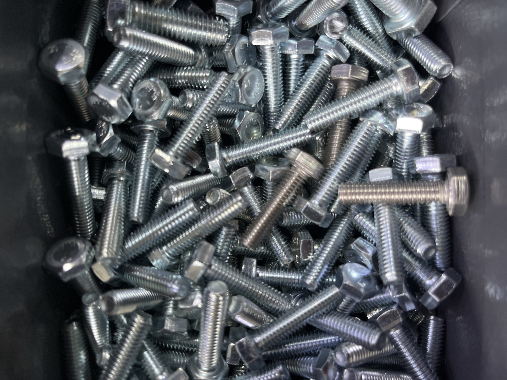 The image shows a close-up, top-down view of a large pile of hex-head bolts. The bolts are made of metal and appear to be silver or galvanized. They are of various sizes and are jumbled together in a chaotic heap. Some bolts are lying flat, while others are propped up at angles. The background is dark and out of focus, suggesting the bolts are contained in a box or bin. There is a single bolt in the foreground that is upright and clearly visible. The overall impression is one of abundance and disarray. There are no people or specific landmarks visible that would allow for the identification of a location in Kyiv, Ukraine.