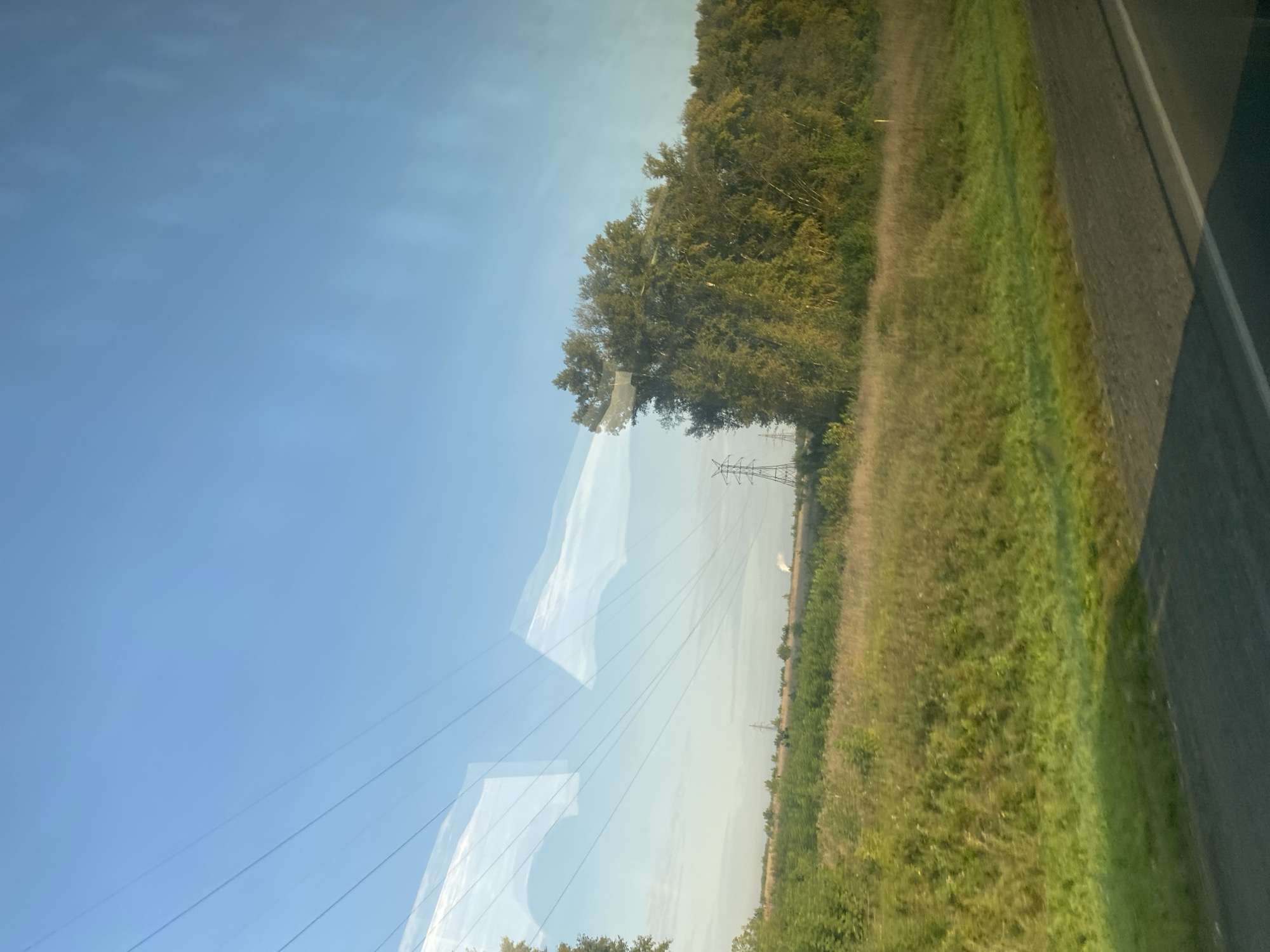The image depicts a rural landscape viewed from a moving vehicle, likely a car, indicated by the road surface in the foreground and the reflections on the window. The scene is dominated by a vast, clear blue sky, with several high-tension power lines stretching across the frame. Below the sky, a grassy field with scattered trees and shrubs occupies the middle ground. In the distance, a pylon structure for power lines is visible, along with what appears to be a factory or industrial facility emitting a plume of smoke or steam. The overall impression is one of a semi-industrialized rural area under a bright sky. The scene does not contain any people, animals, or specific identifiable landmarks that would pinpoint its exact location within Kemerovo, Russia.